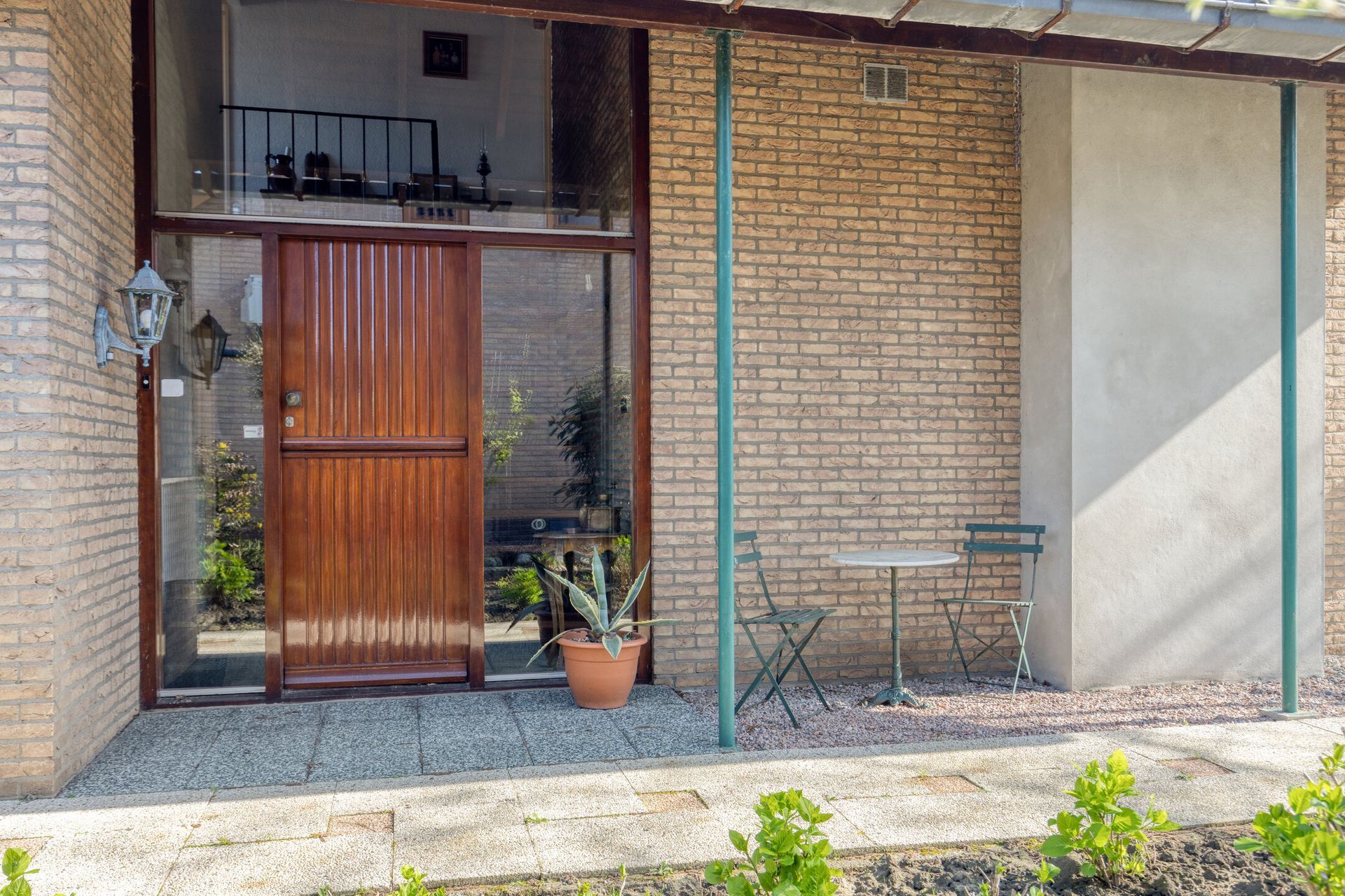 Veranda met houten deur, bakstenen muren, potplant en een klein tafeltje met stoelen onder een schaduwrijk afdak.