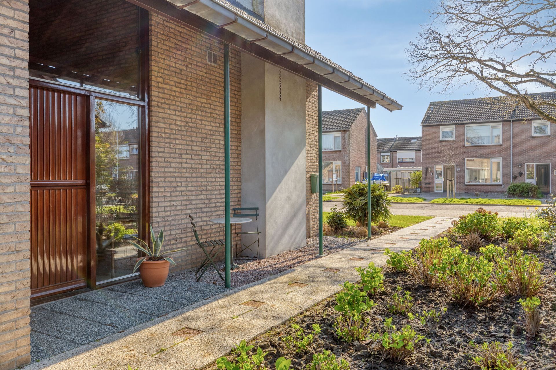 Ingang van een bakstenen huis met houten deur, potplant en tuinpad dat naar de appartementengebouwen leidt.