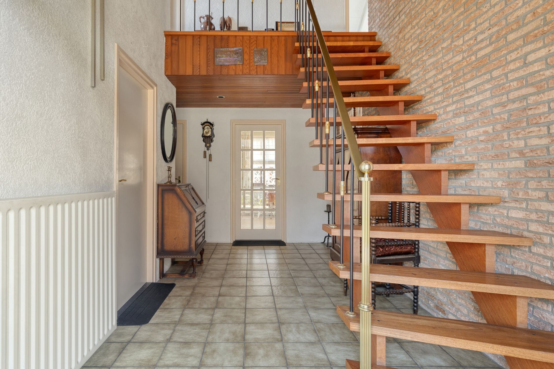Trappenhal met tegelvloer, stenen muur, witte voordeur en houten balustrade naar de zolder.