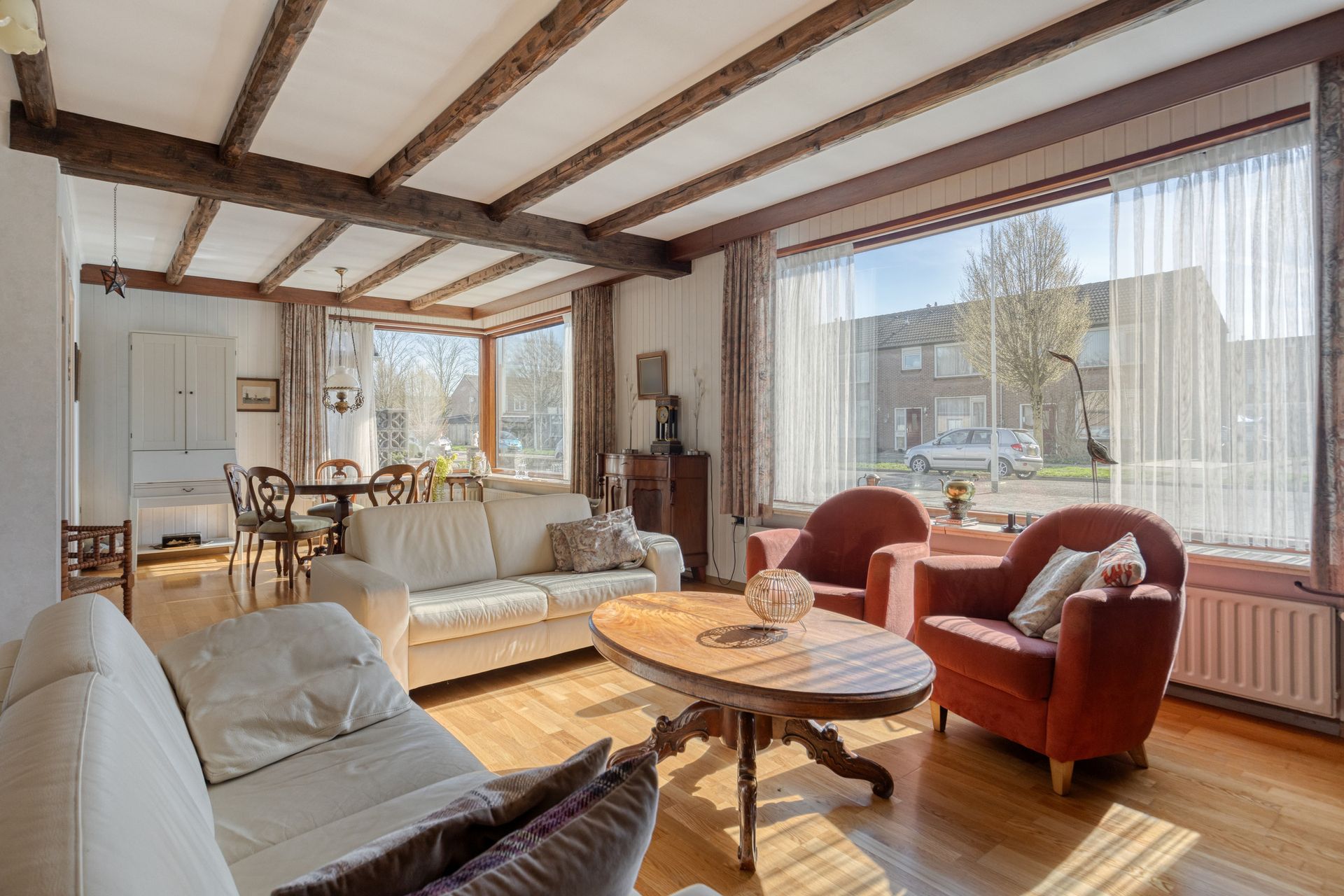 Lichte woonkamer met houten balken, grote ramen, witte bank, rode stoelen en een ronde salontafel.