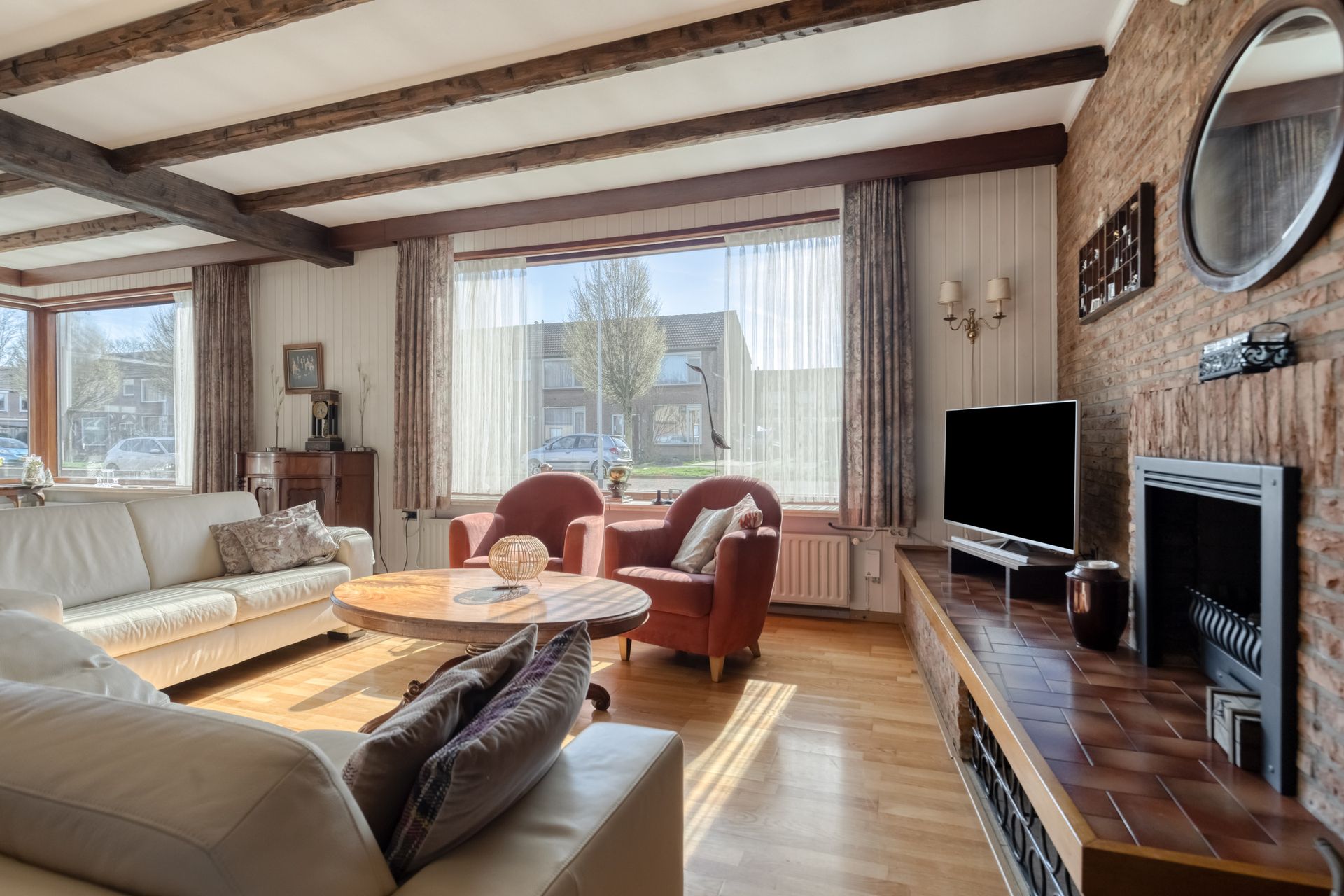 Gezellige woonkamer met beige banken, fauteuils, grote ramen en een open haard met tv.