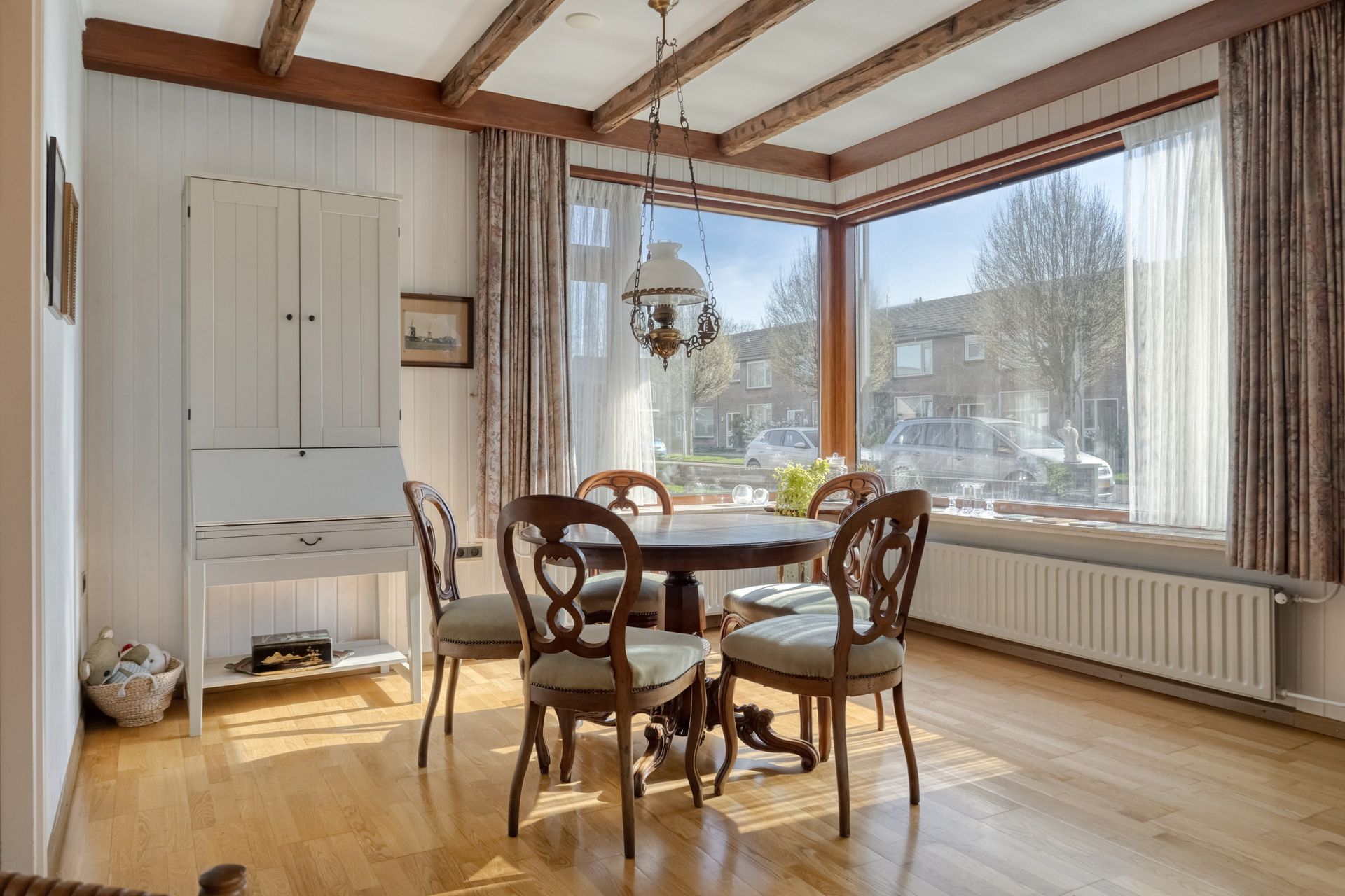 Lichte eetkamer met houten tafel en stoelen, grote hoekramen en zonlicht op de houten vloer.