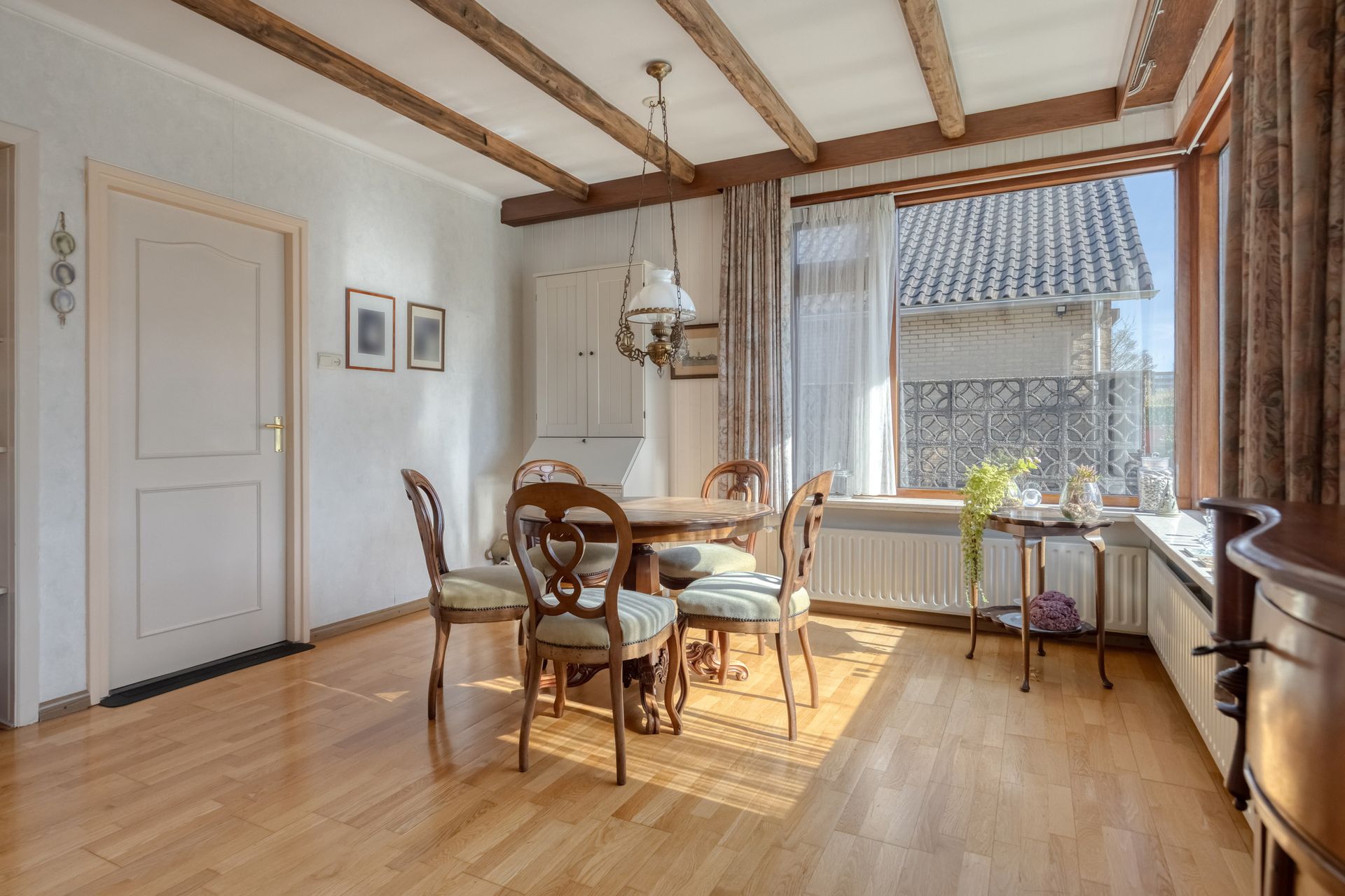 Lichte eetkamer met houten balken, tafel en stoelen, en een zonnig raam met gordijnen.