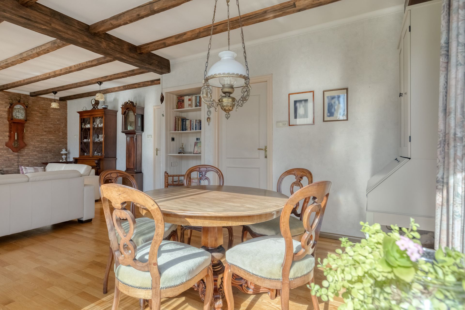 Lichte eetkamer met houten balken, ronde tafel, stoelen, witte kasten en een hangende kroonluchter.