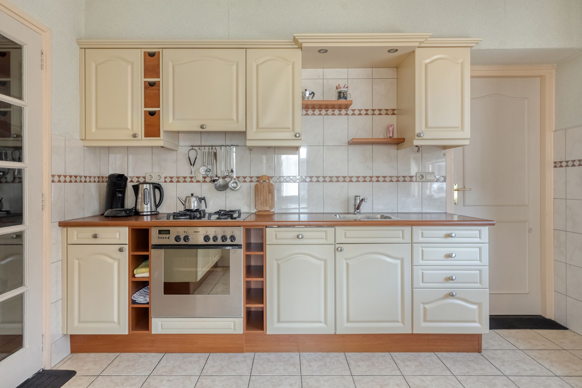 Crèmekleurige keuken met kasten, oven, spoelbak, aanrechtblad en tegelvloer.