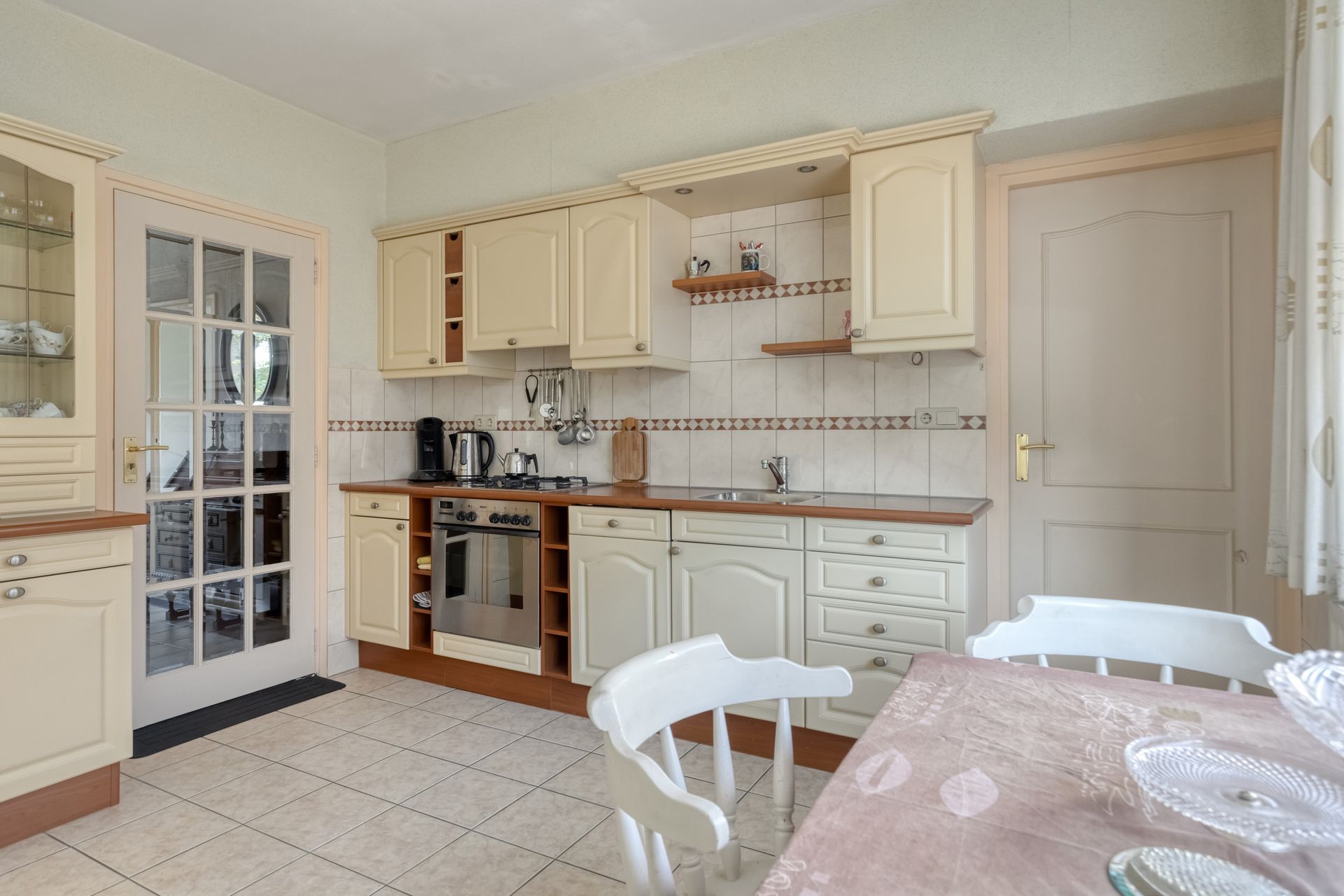 Lichte keuken met crèmekleurige kasten, tegelvloer, witte deur en een tafel met stoelen op de voorgrond.