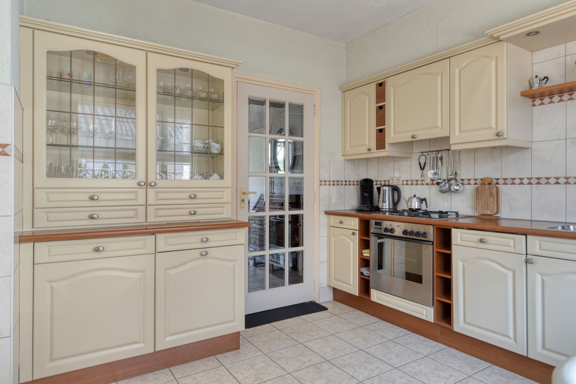 Lichte crèmekleurige keuken met kastjes met glazen deuren, witte apparaten, houten afwerking en een tegelvloer.