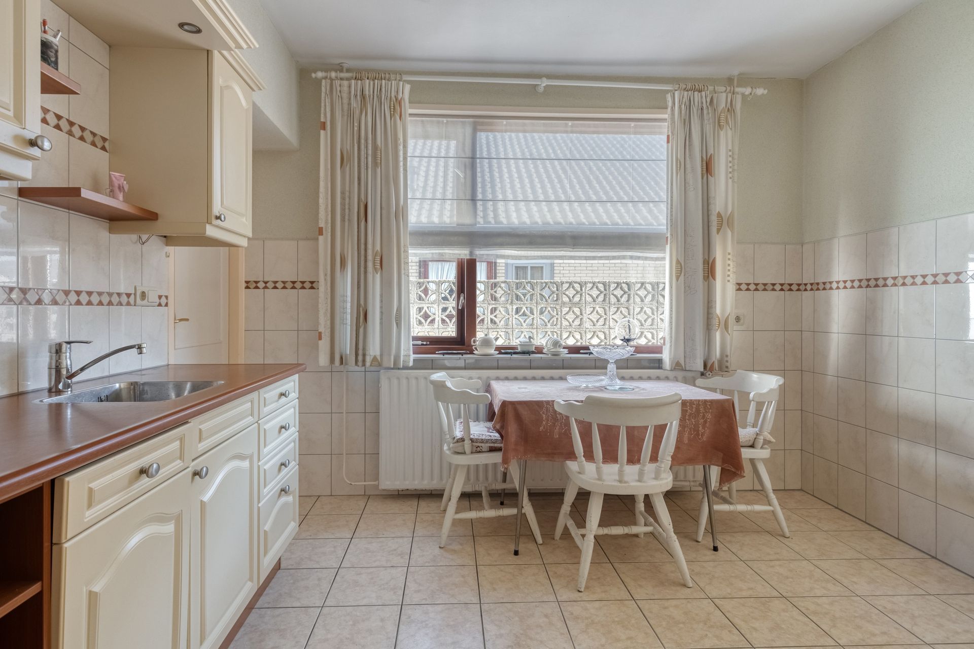 Lichte keuken met witte kasten, betegelde muren, een kleine eettafel en een raam met gordijnen.