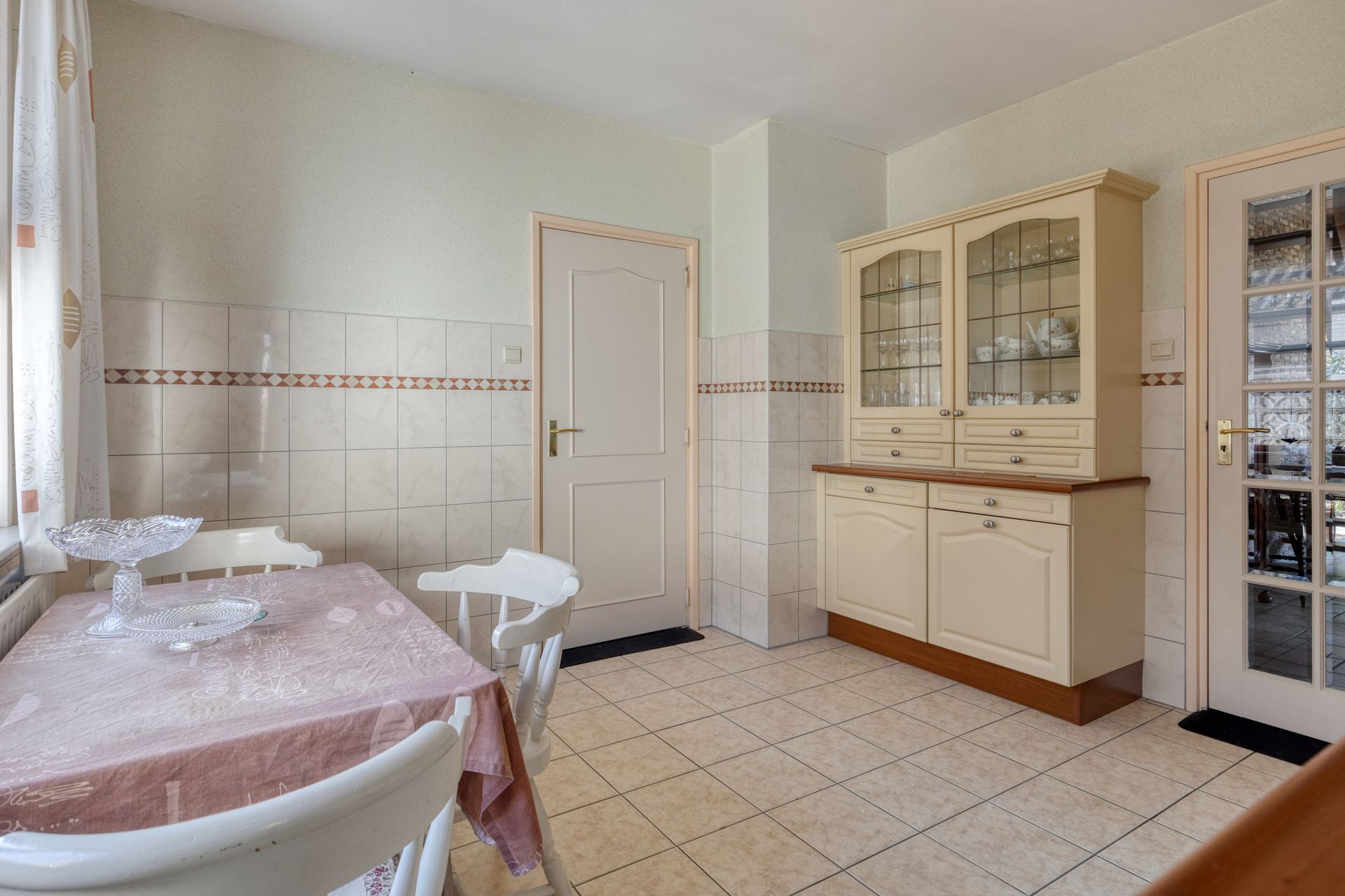 Lichte keuken met witte kasten, tegelvloer en een tafel met een roze tafelkleed bij een gesloten deur.