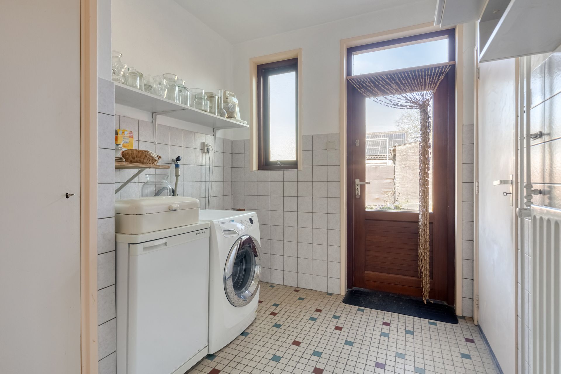 Kleine wasruimte met wasmachine, wasbak, planken, betegelde muren en een open houten deur naar buiten.
