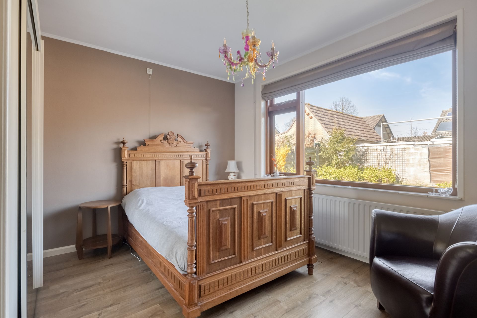 Lichte slaapkamer met sierlijk houten bed, groot raam, zwarte fauteuil en kroonluchter.