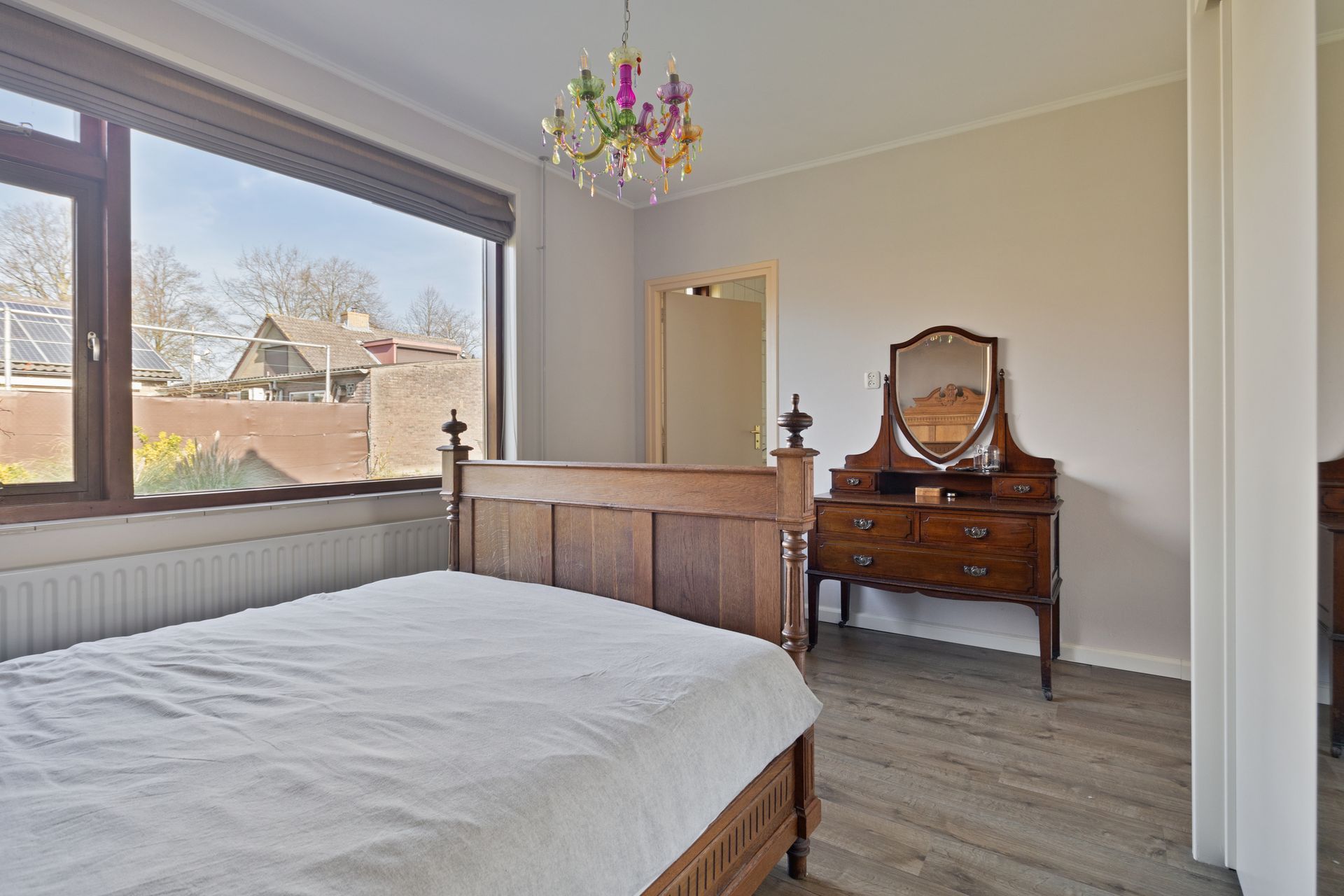 Lichte slaapkamer met een bed, commode en een groot raam met uitzicht op bomen en daken.