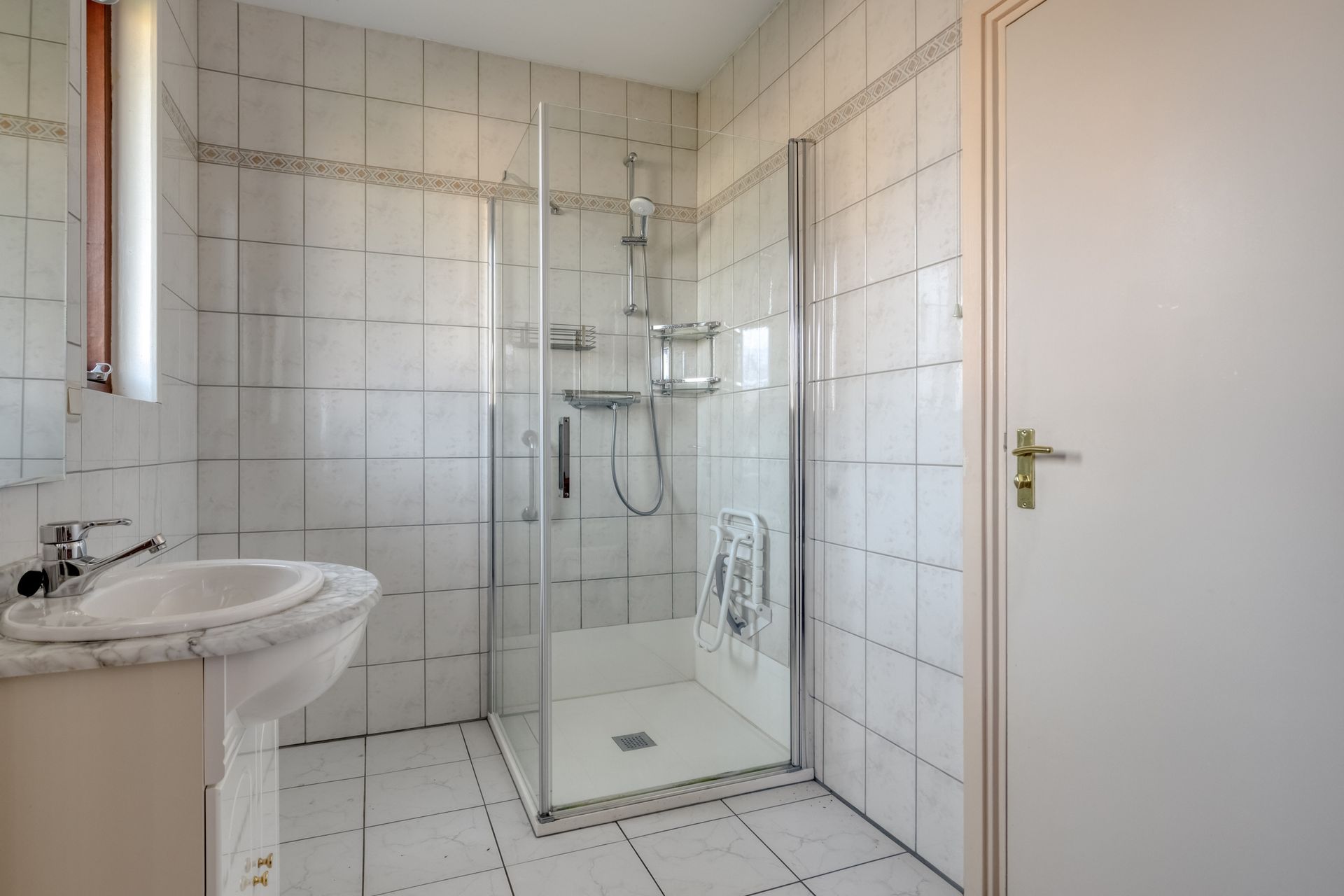 Kleine betegelde badkamer met glazen douchewand, wastafel en witte deur.