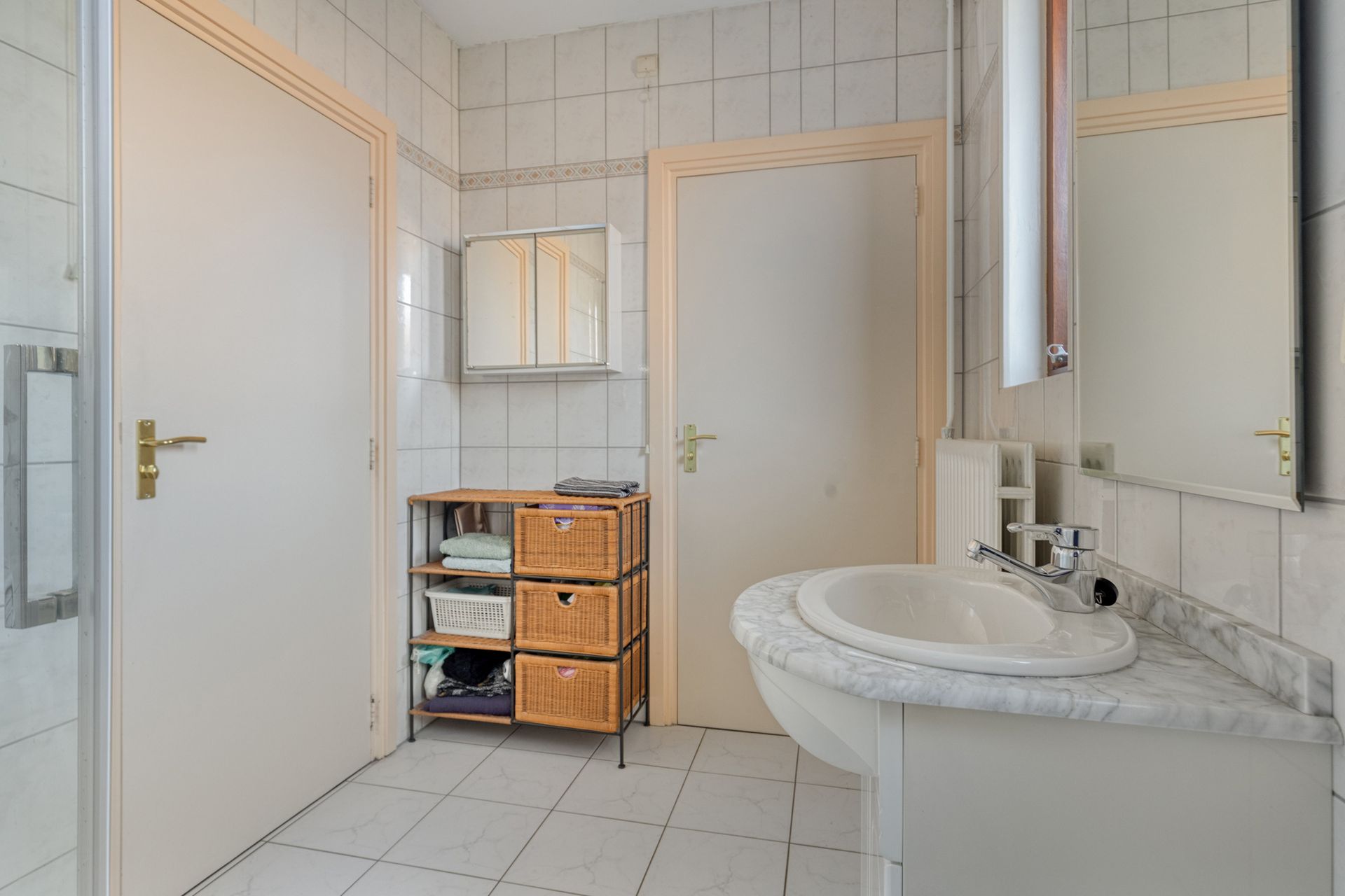 Lichte badkamer met witte tegels, twee deuren, een rieten kast en een marmeren wastafel met spiegel.