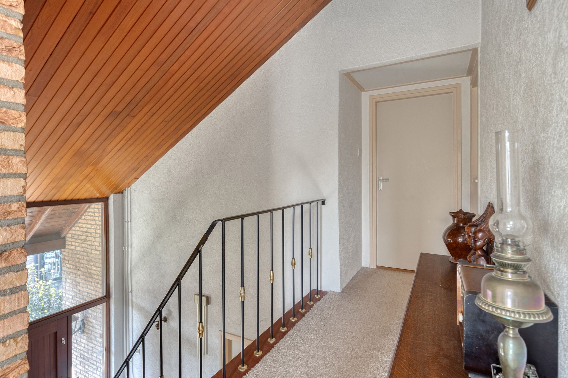 Trappenhuis met schuin houten plafond, zwarte leuning en een witte haldeur.