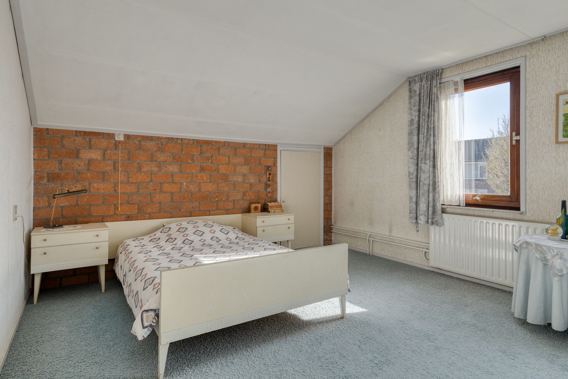 Lichte slaapkamer met een wit bed, een accentmuur van baksteen en een raam met gordijnen.