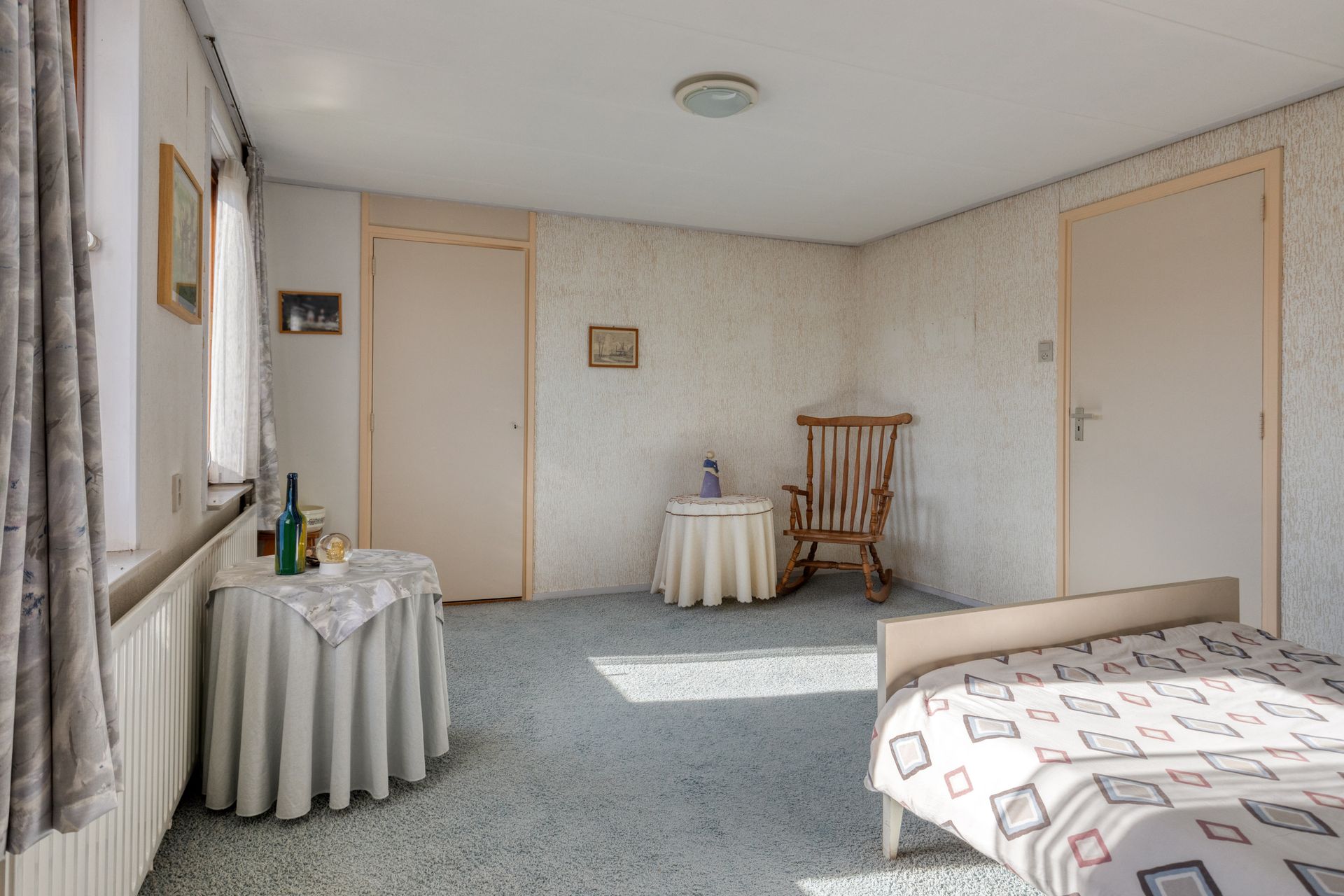 Kleine slaapkamer met twee eenpersoonsbedden, licht behang, vloerbedekking en een raam waar zonlicht doorheen komt.