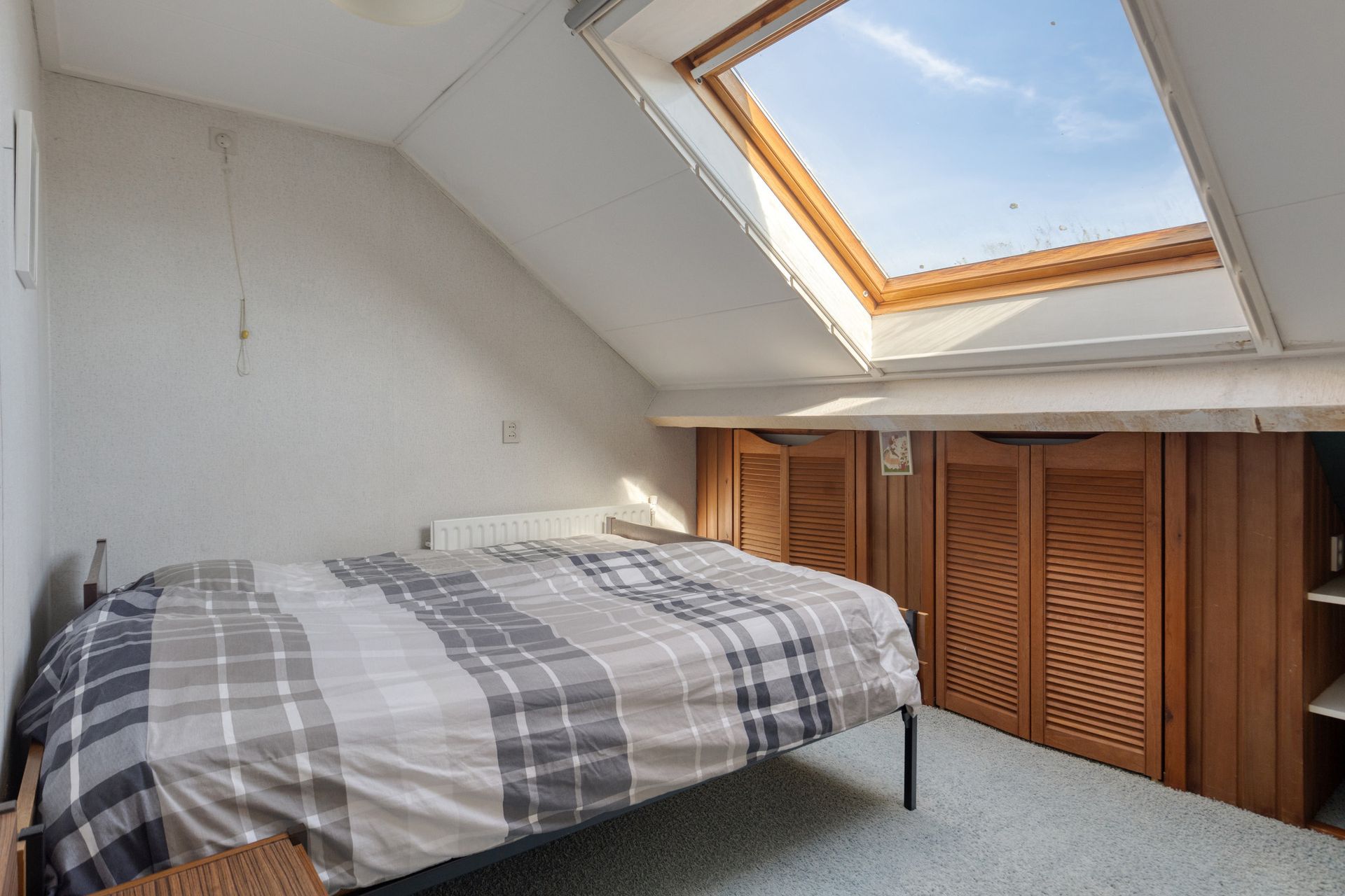 Slaapkamer met bed onder een dakraam en schuin plafond, voorzien van houten kastdeuren en grijs tapijt.