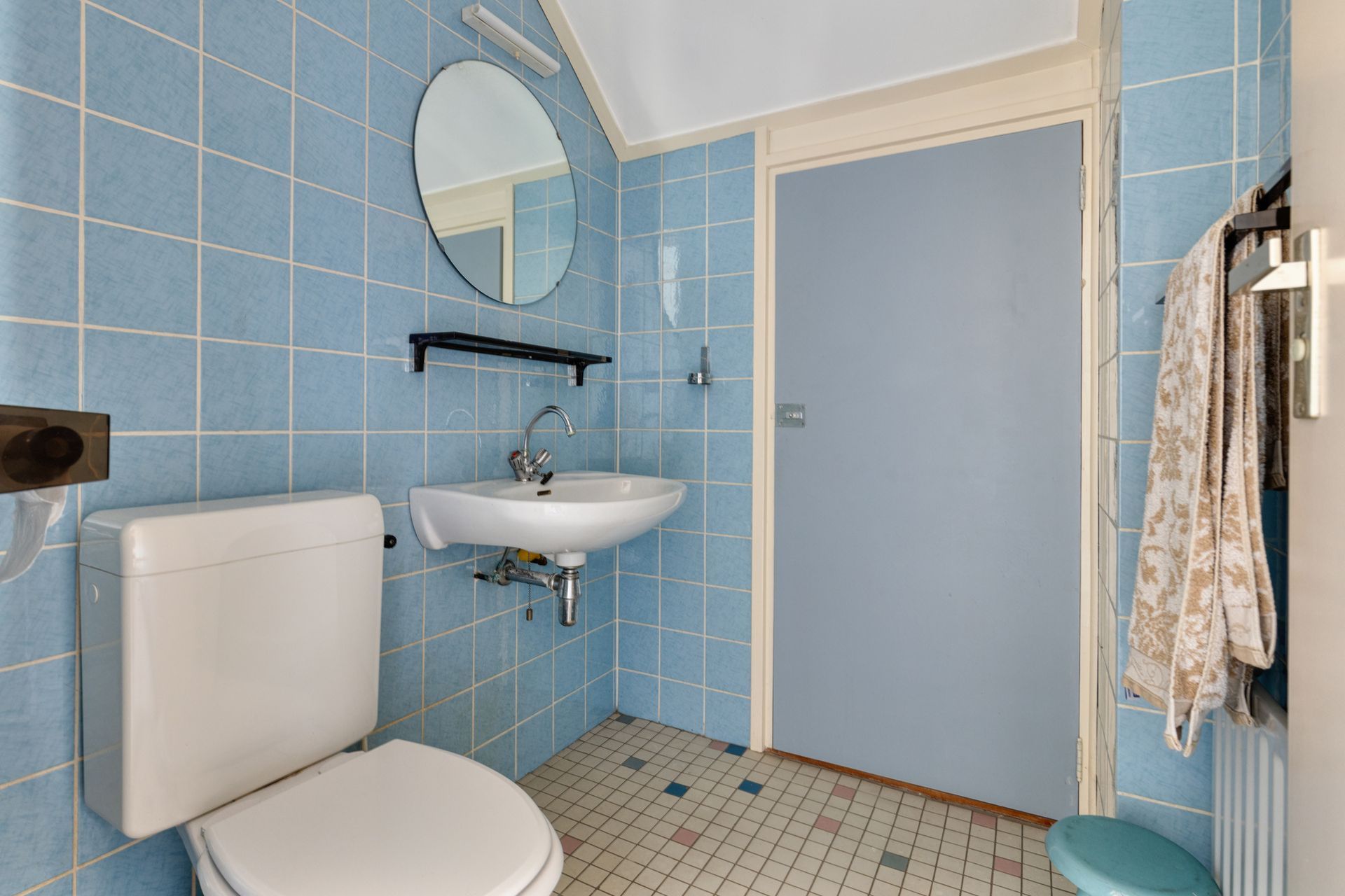 Badkamer met blauwe tegels, toilet, wastafel, ronde spiegel, douchedeur en handdoekrek.