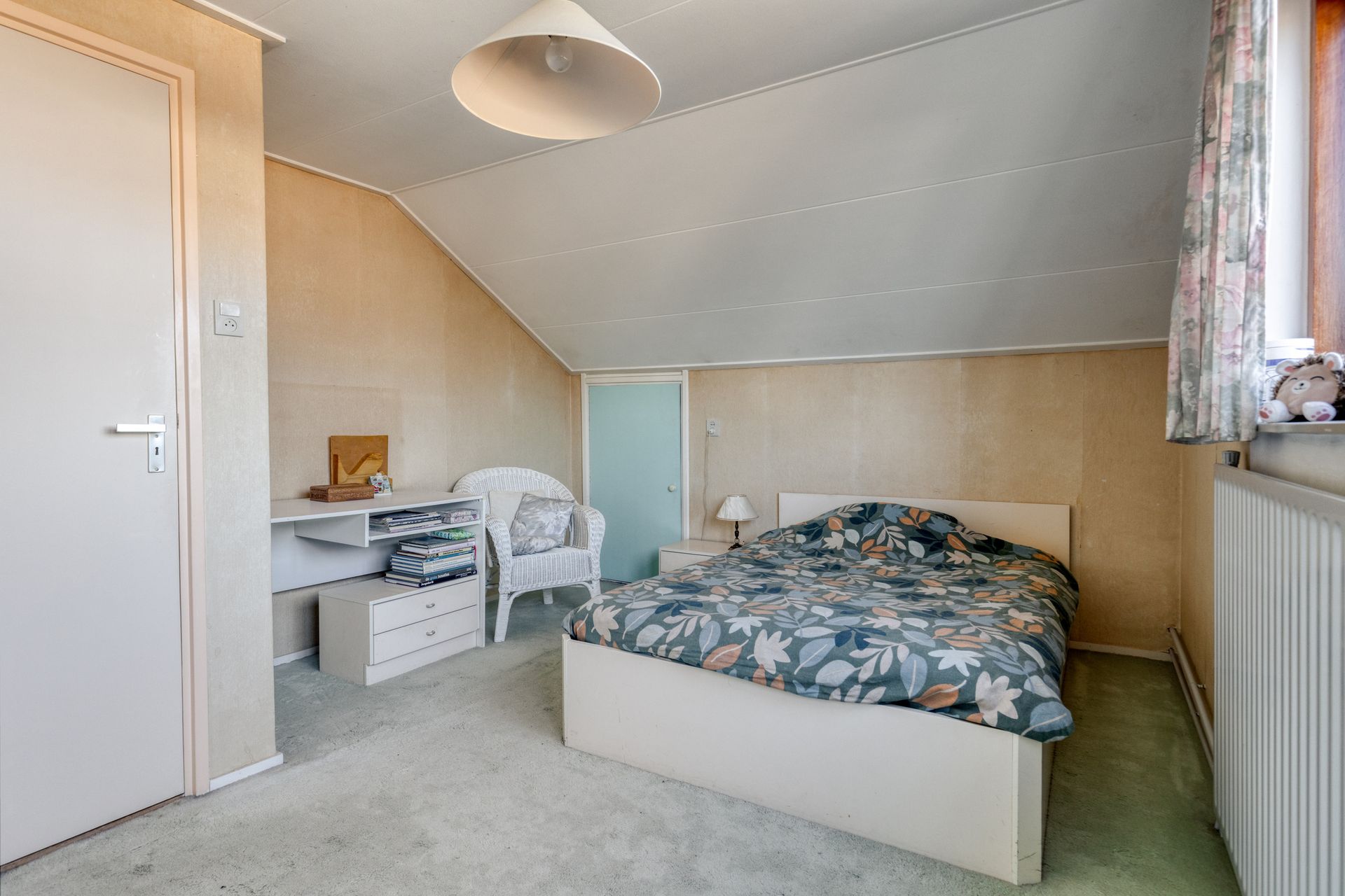 Kleine zolderkamer met een bed, nachtkastje, bureau en schuin plafond in beige tinten.
