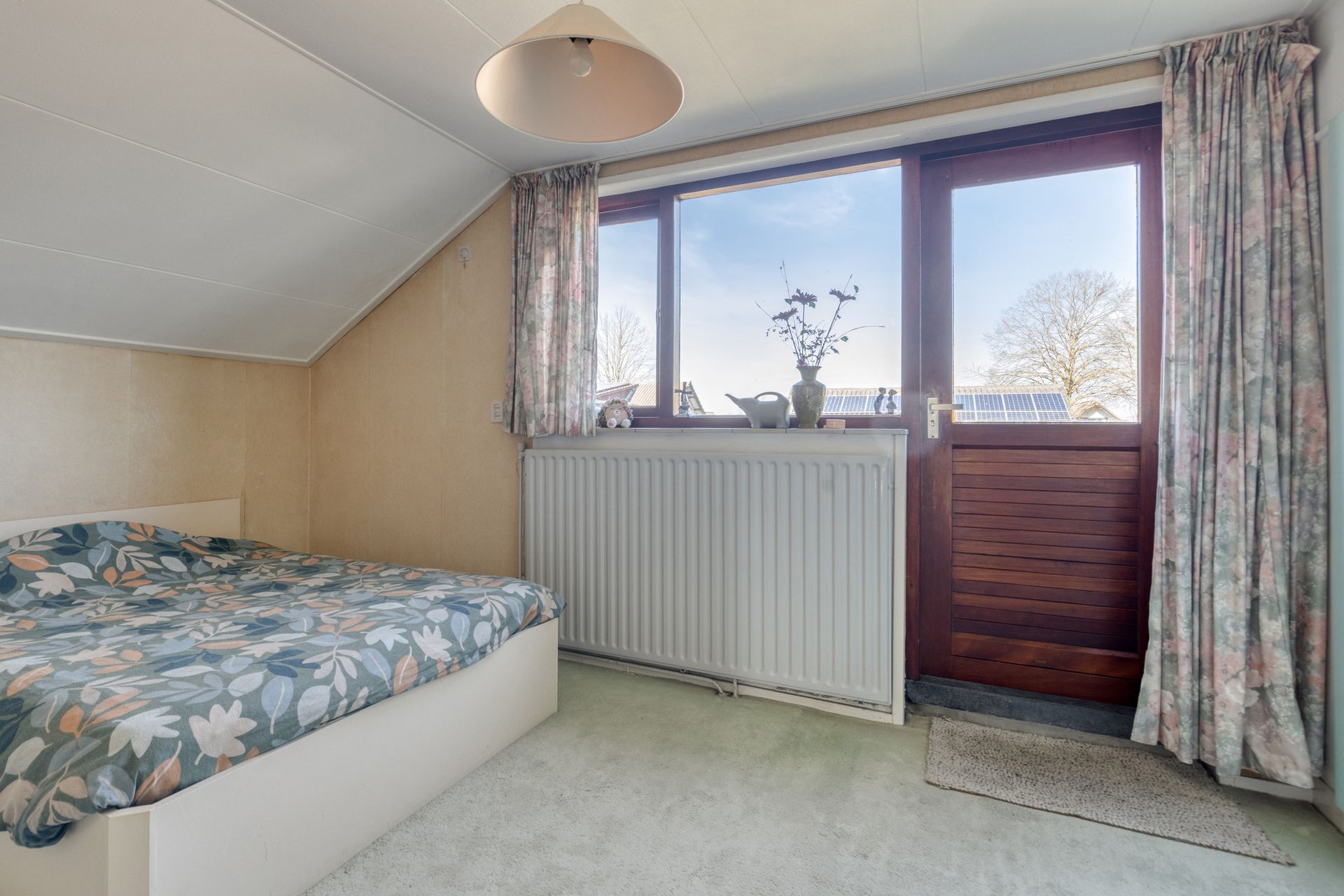 Kleine slaapkamer met bloemensprei, schuin plafond, raam, radiator en glazen deur met gordijnen.