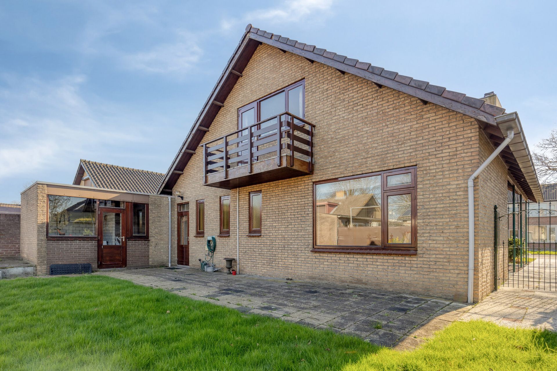 Bakstenen huis met een klein balkon, terras en groen gazon onder een blauwe hemel.