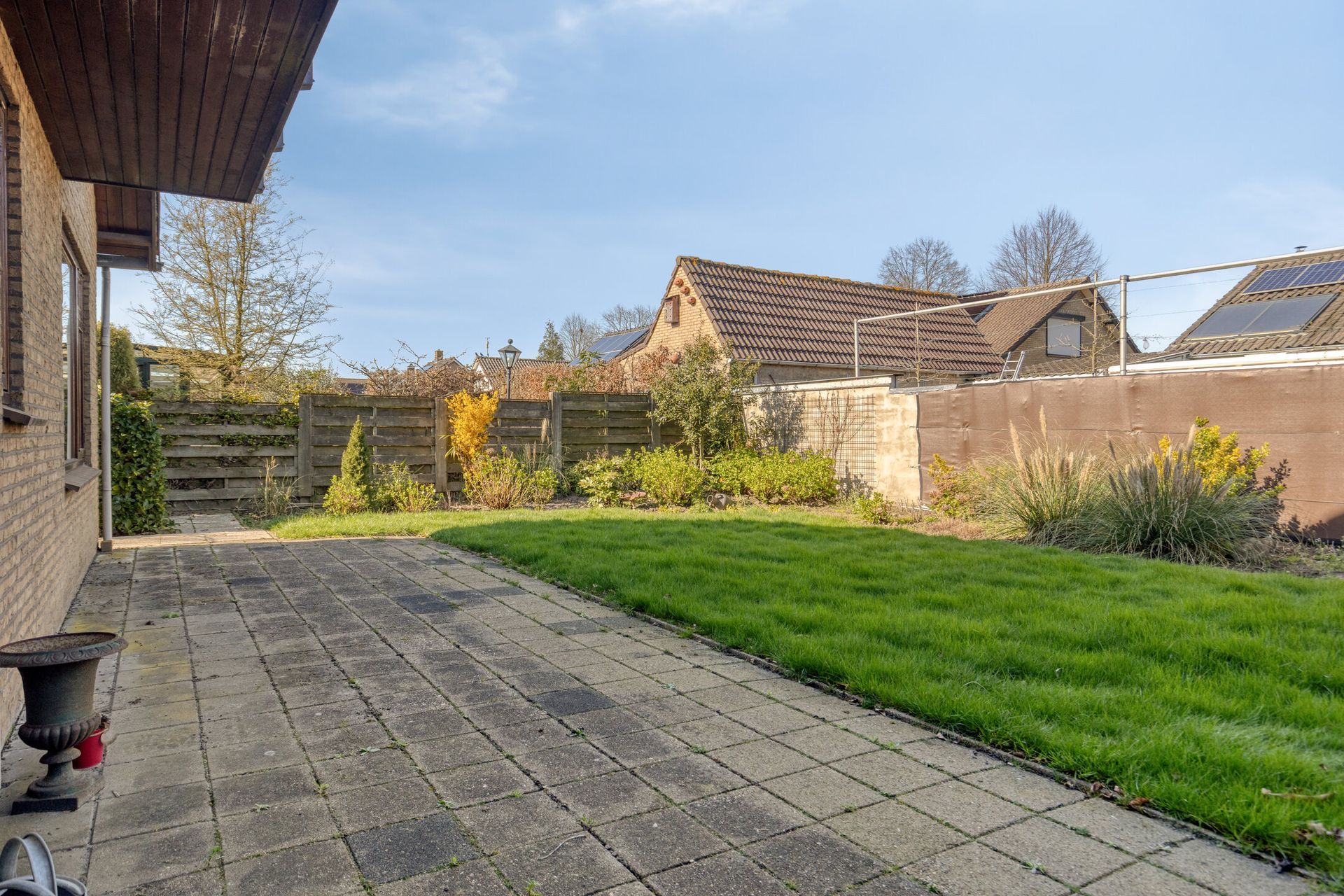 Terras in de achtertuin met bestrating, groen gazon en uitzicht op de huizen van de buren onder een blauwe hemel.