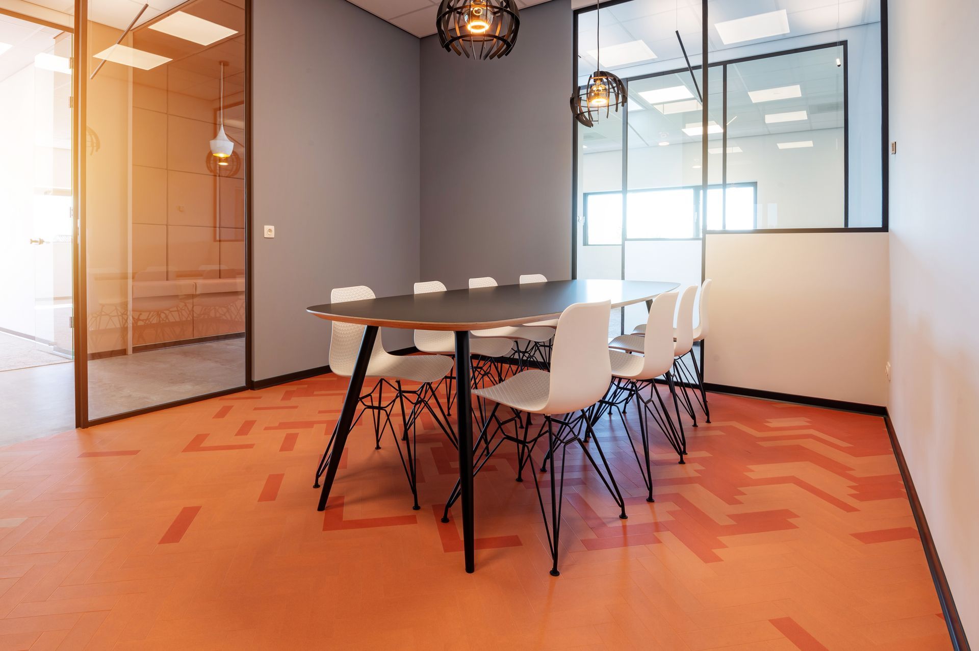 Vergaderruimte met ovale tafel, witte stoelen, oranje vloer met patroon, glazen wanden en hanglampen.