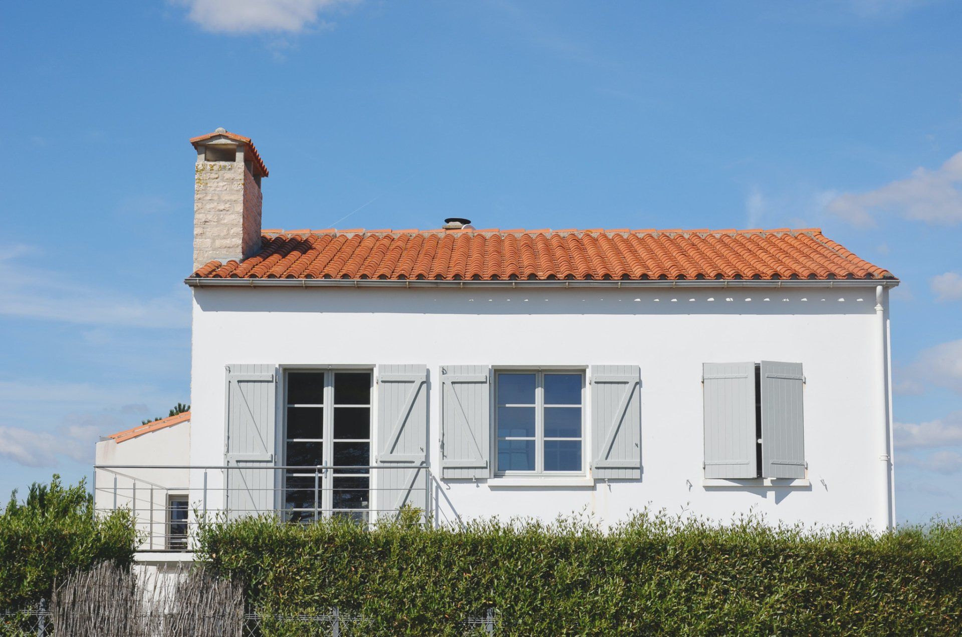 Een wit huis met een oranje pannendak, grijze luiken en een bakstenen schoorsteen tegen een helderblauwe hemel.