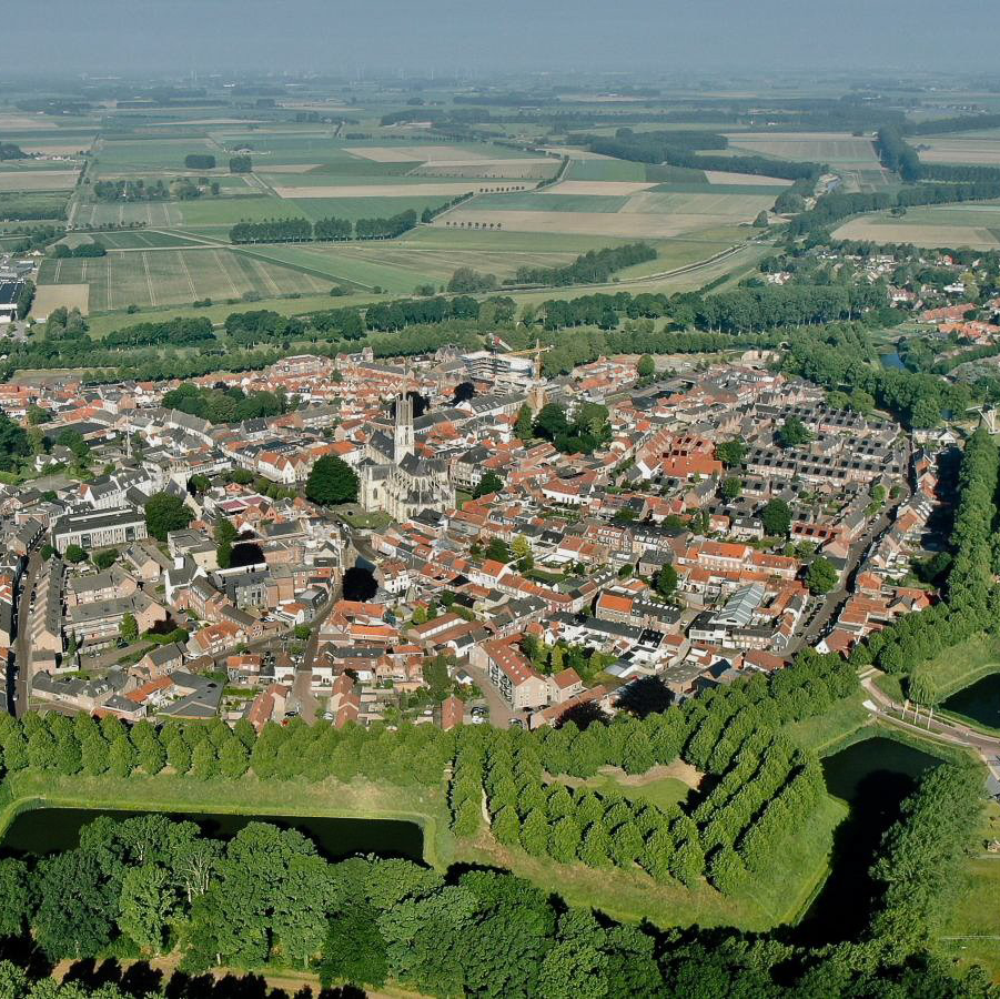 Luchtfoto van een ommuurd Nederlands stadje met rode daken, een kerktoren, omgeven door een groene gracht en landbouwgrond.