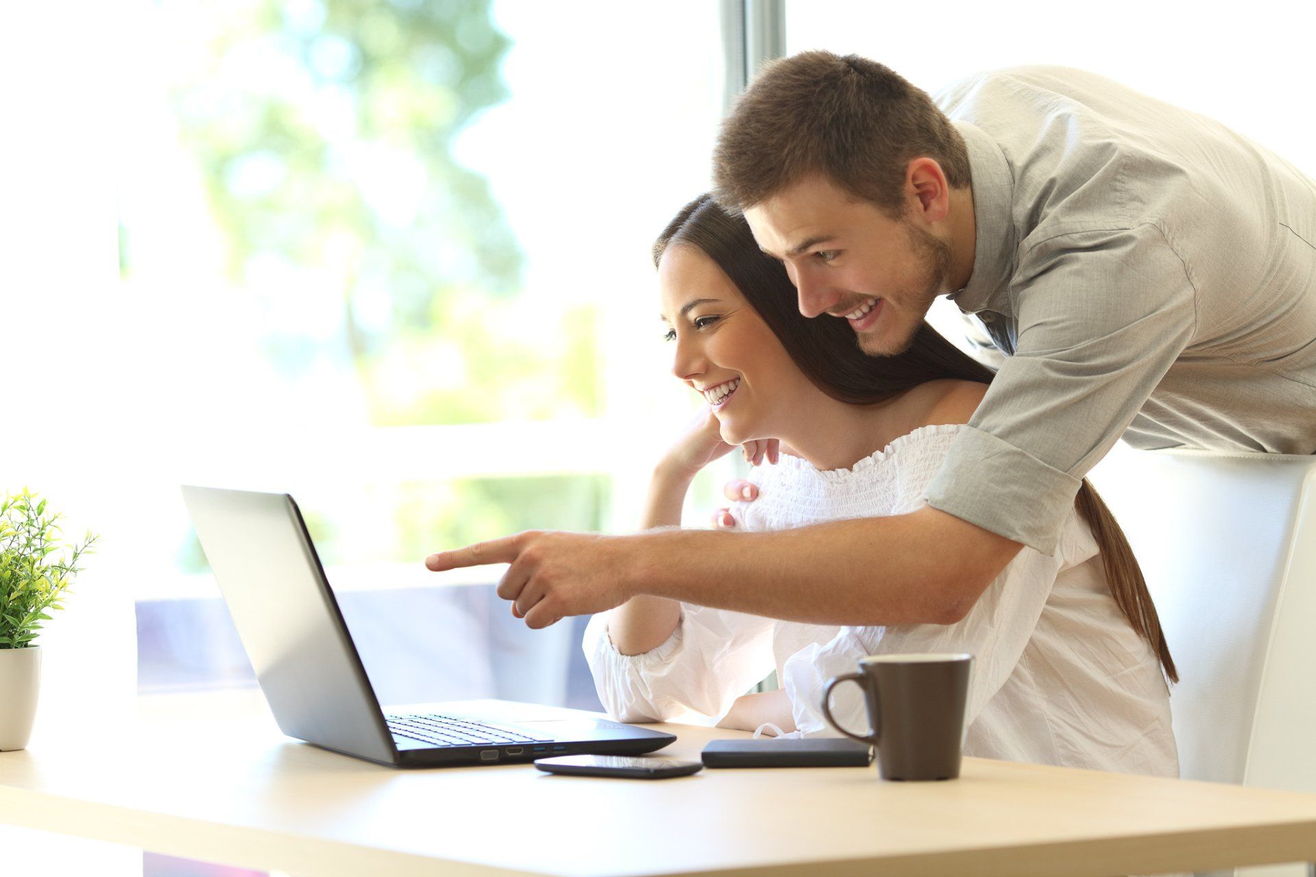 Vrouw en man kijken naar een laptop en glimlachen. De man wijst naar het scherm. Tafel, raam, mok.