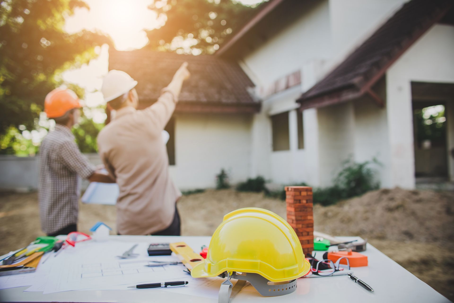 Deux ouvriers du bâtiment examinent des plans devant une maison, des outils sont posés sur une table au premier plan.