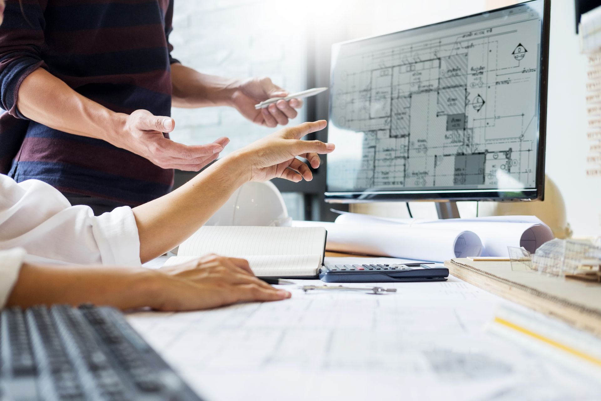 Deux personnes examinent des plans architecturaux sur un écran d'ordinateur et sur papier, en discutant des détails.