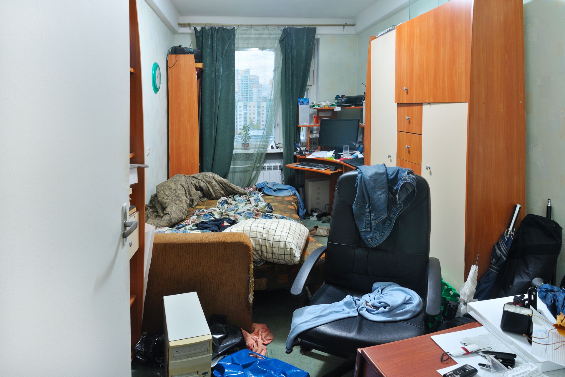 Chambre encombrée avec un lit, un bureau, des armoires et une chaise. Des vêtements et divers objets sont éparpillés