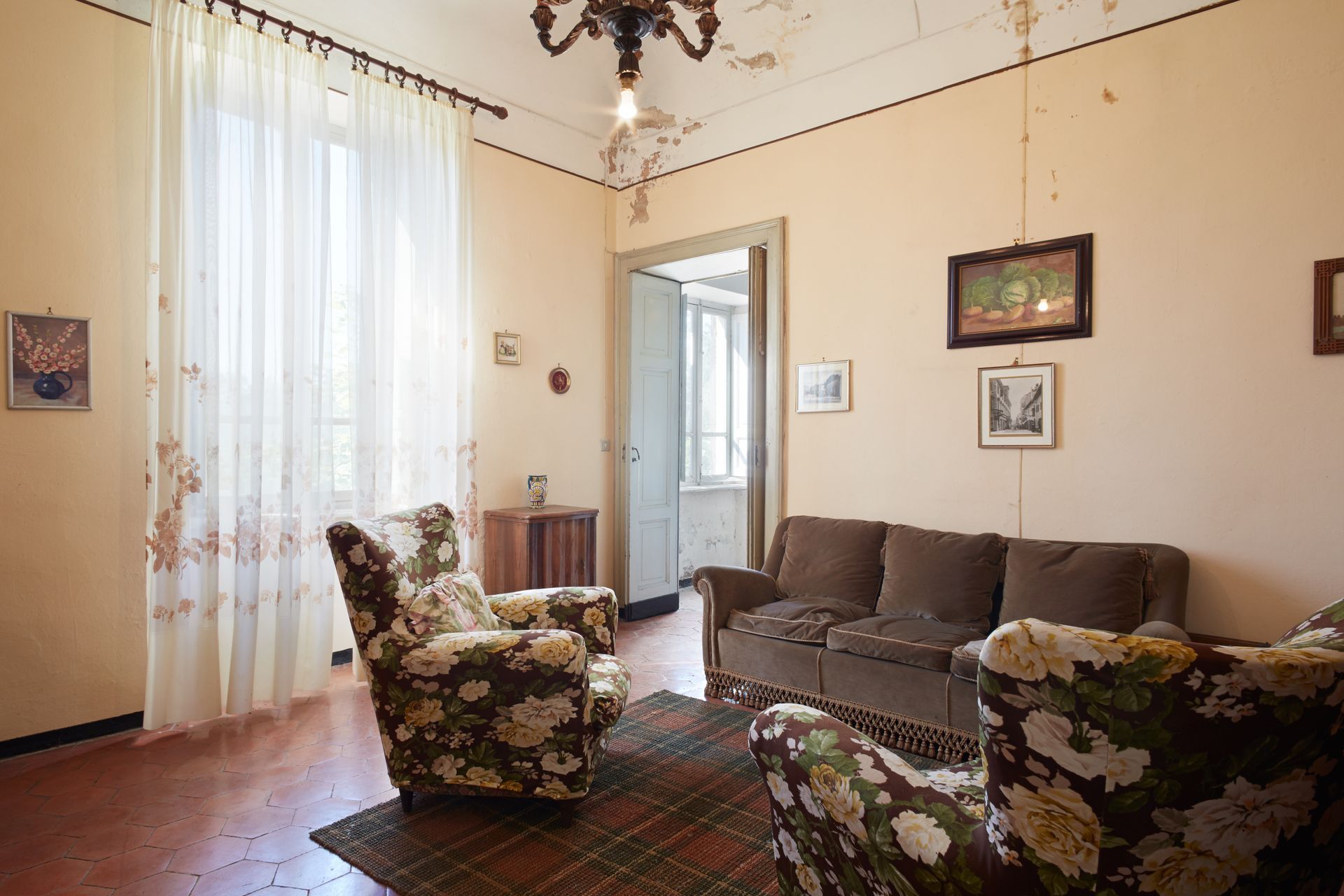 Salon avec fauteuils à motifs floraux et canapé marron, rideaux vaporeux et œuvres d'art aux murs.