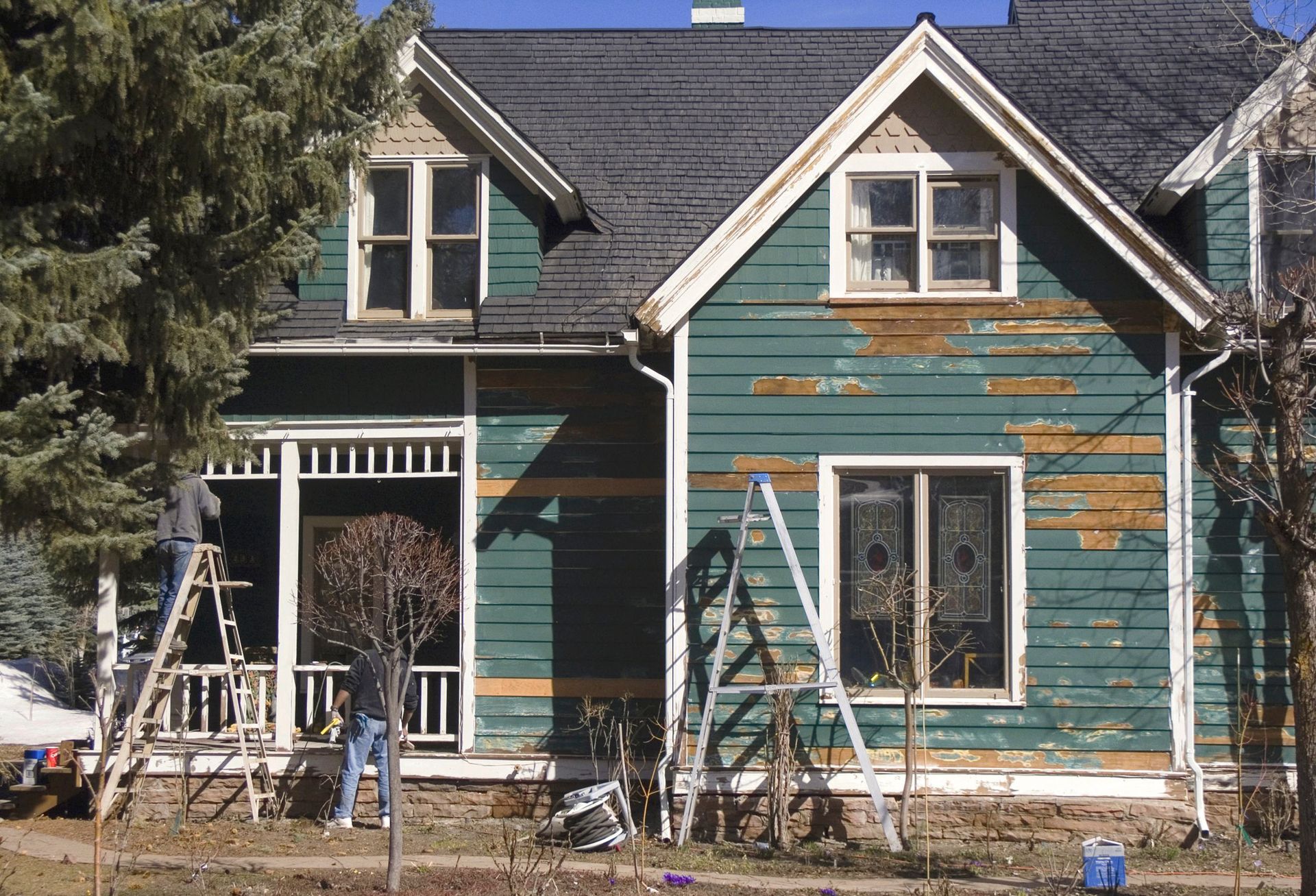 Maison à la peinture turquoise écaillée, en cours de rénovation, avec des échelles et des arbres.