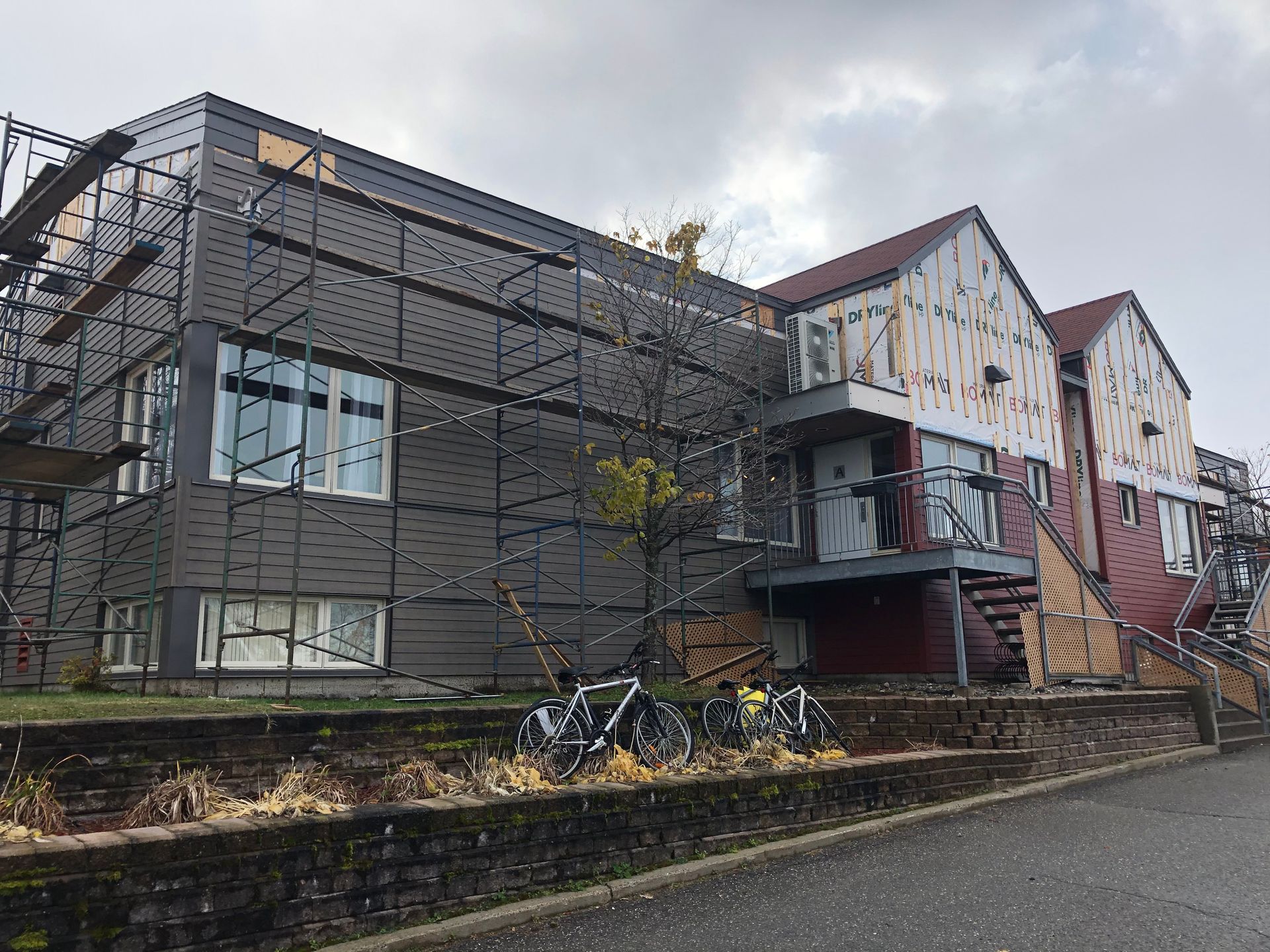 Bâtiment en rénovation avec échafaudages, bardage gris foncé et sections rouges. Vélos garés à l'extérieur.