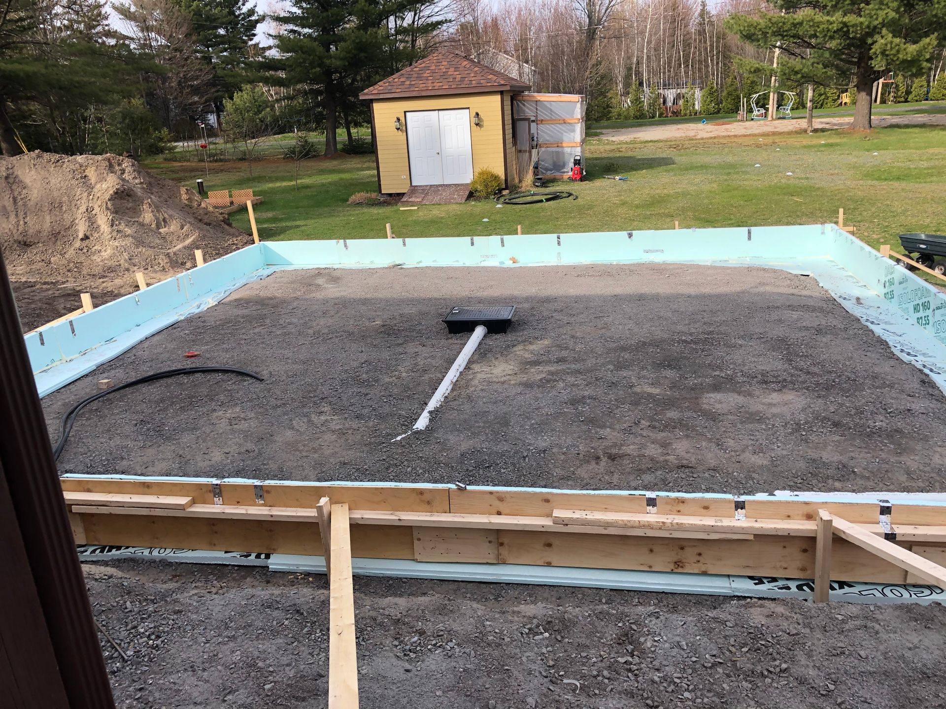 Chantier de construction avec gravier à l'intérieur d'une structure en bois et en mousse bleue ; hangar en arrière-plan.