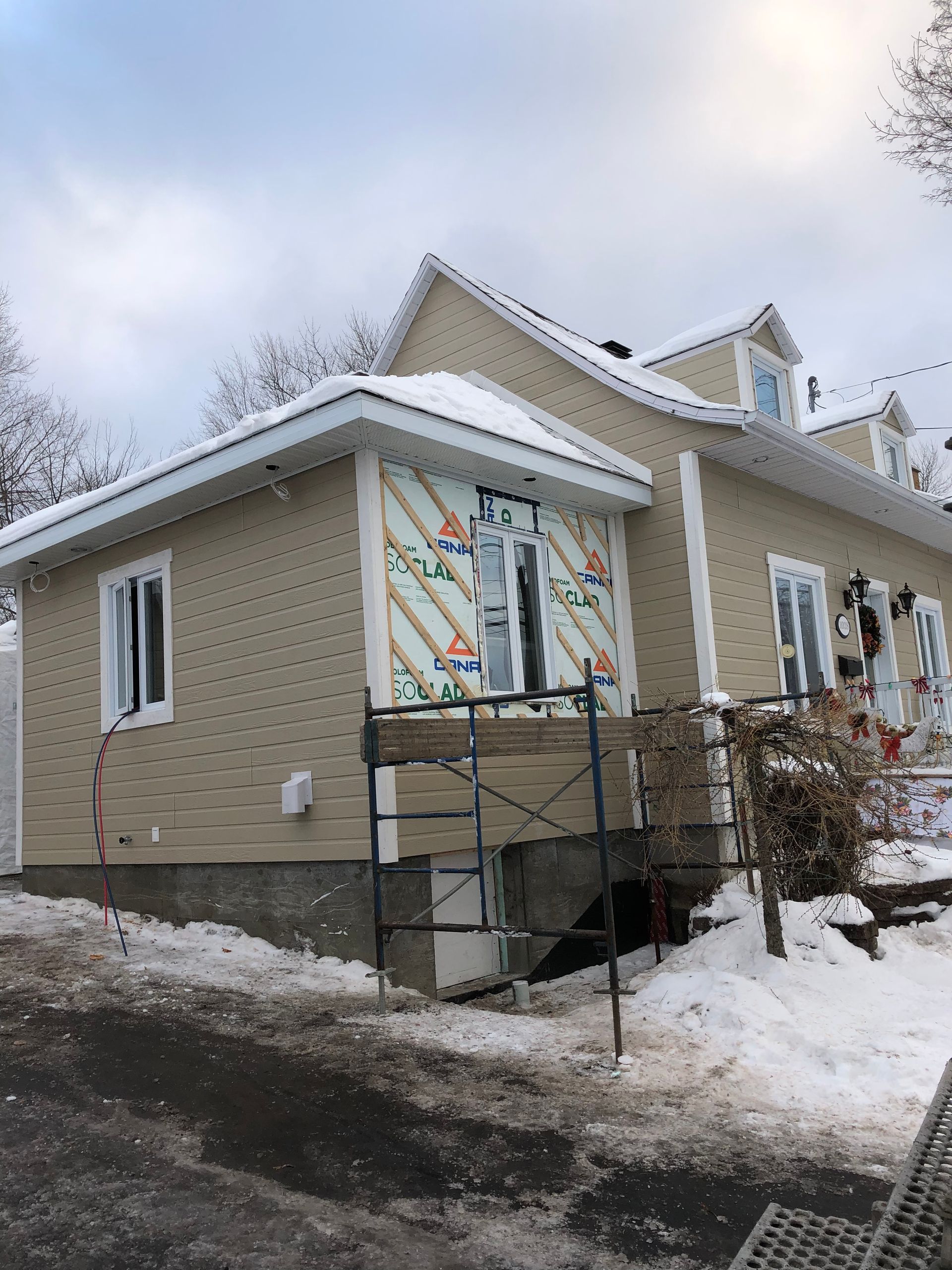 Maison beige en construction, avec isolation apparente autour d'une fenêtre, dans un décor enneigé.