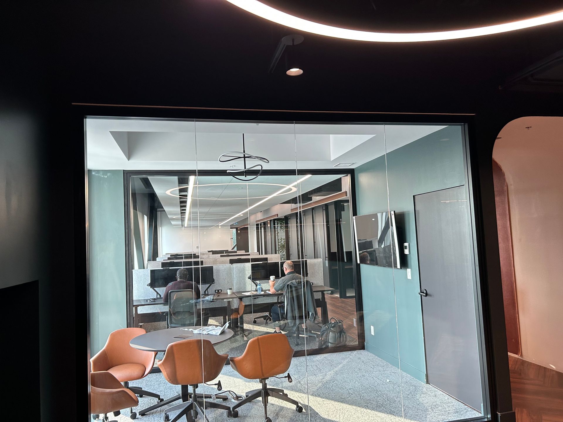 Intérieur de bureau : Vue à travers une porte vitrée d'un espace de travail ouvert avec bureaux, chaises et écrans ;