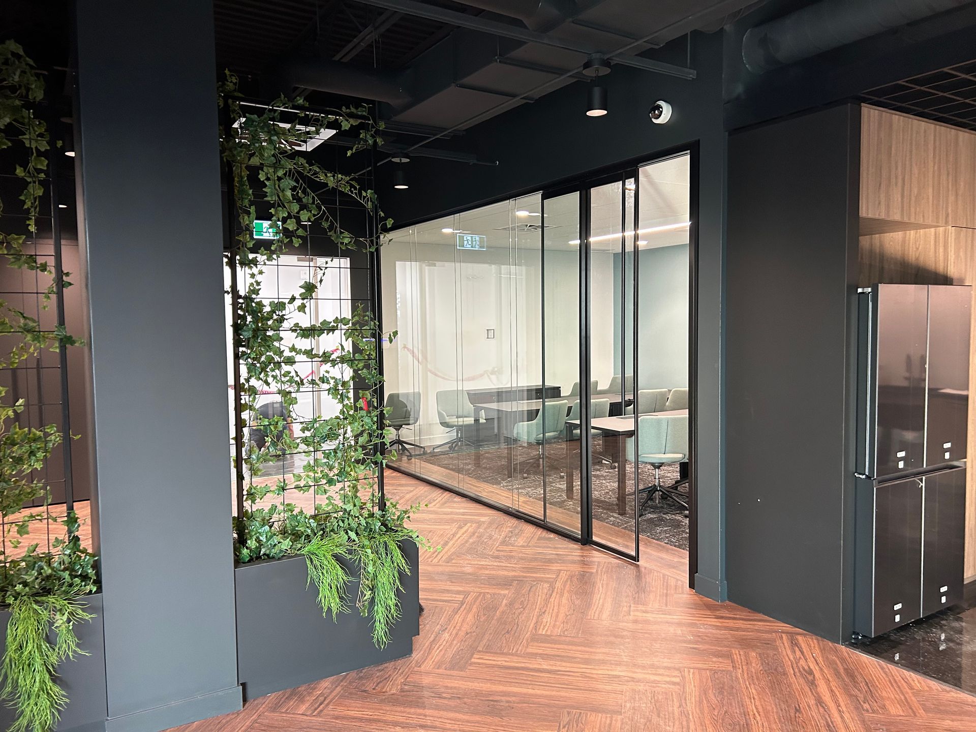 Vue intérieure : un couloir de bureau moderne avec une salle de réunion aux parois vitrées. Plantes vertes et sol en parquet.