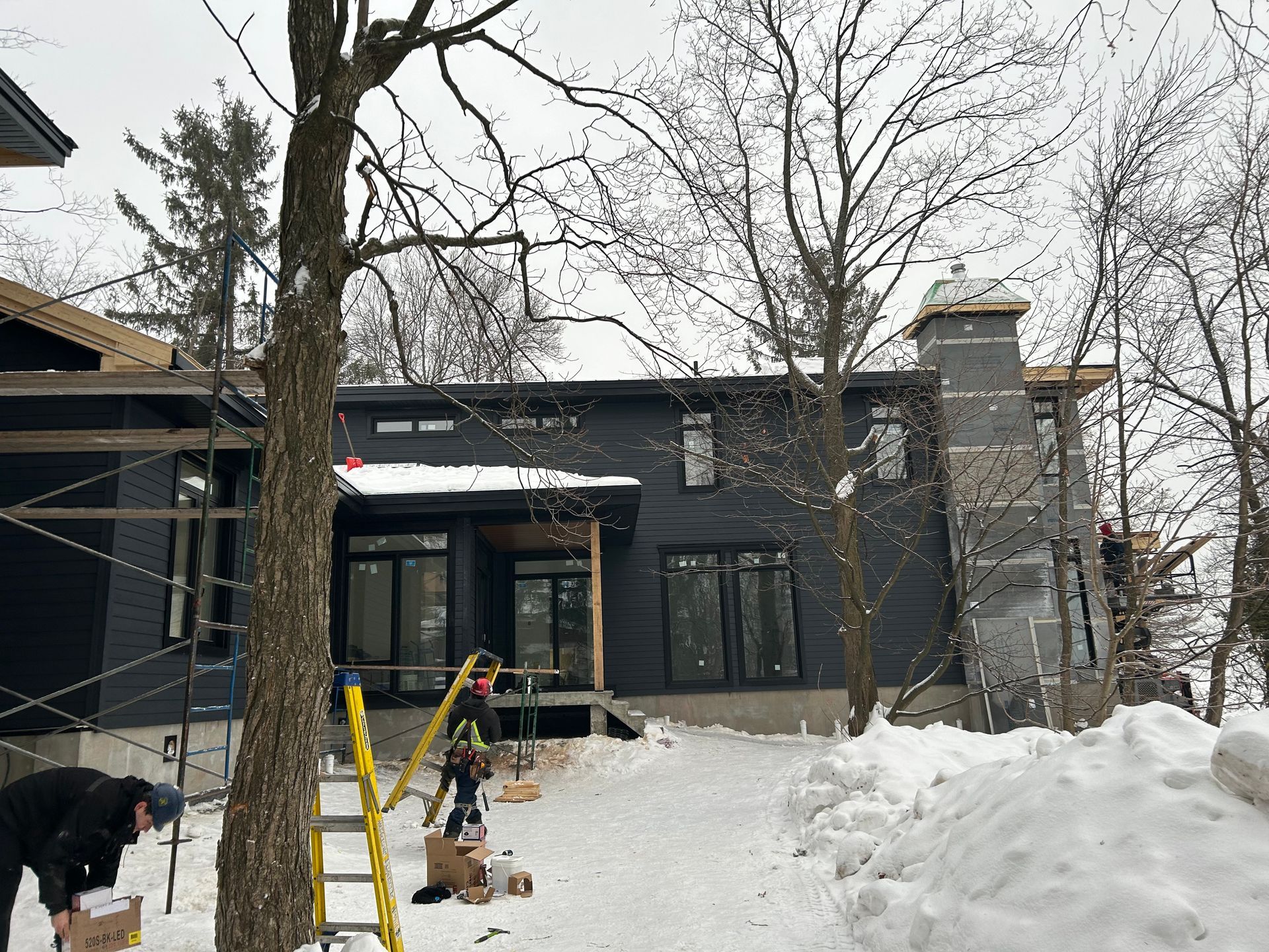 Chantier : maison sombre aux grandes fenêtres, cheminée, neige. Ouvriers et échelles présents. Arbres tout autour.