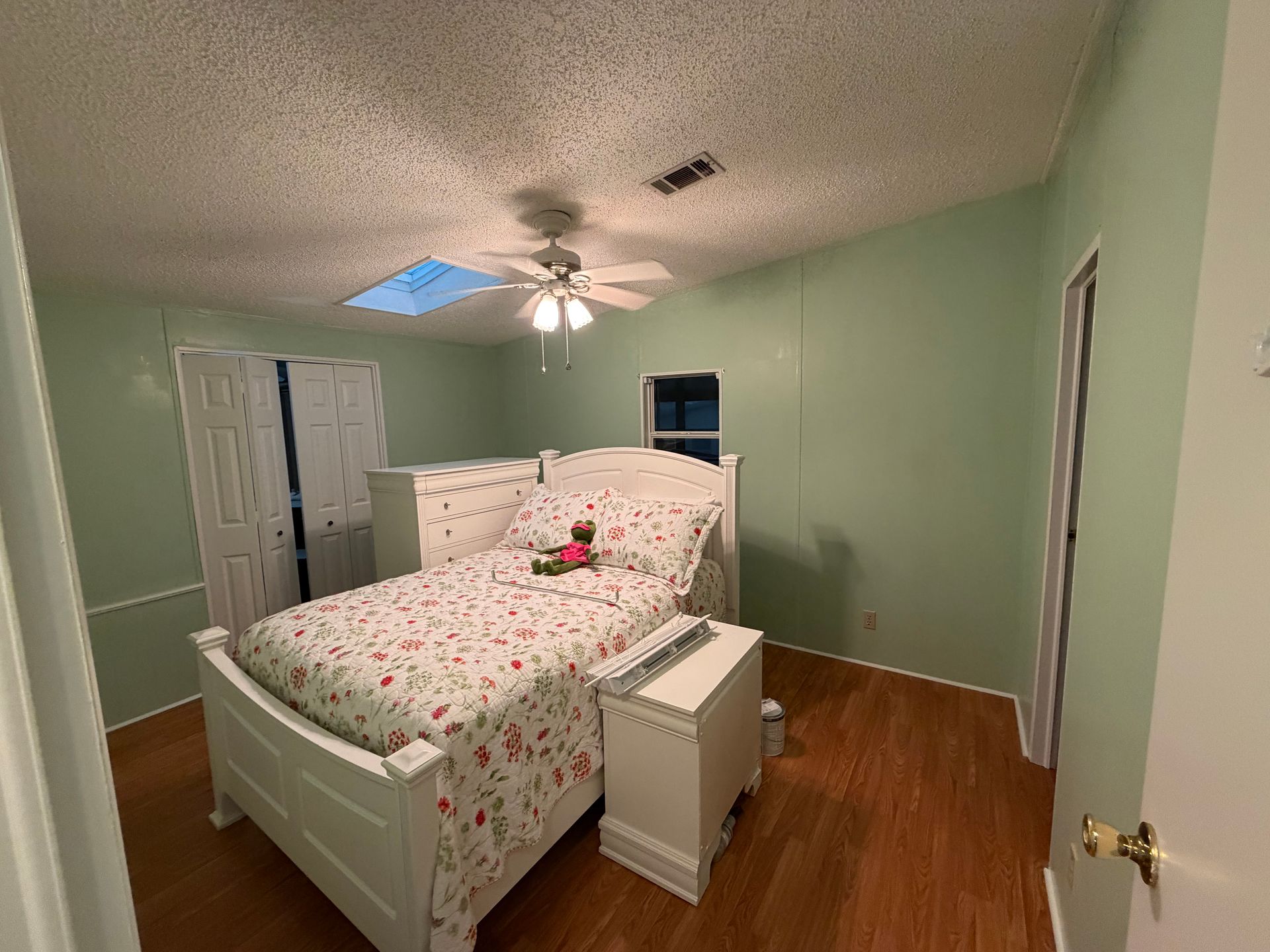 Small bedroom with green walls, white floral bedspread, dresser, and ceiling fan.