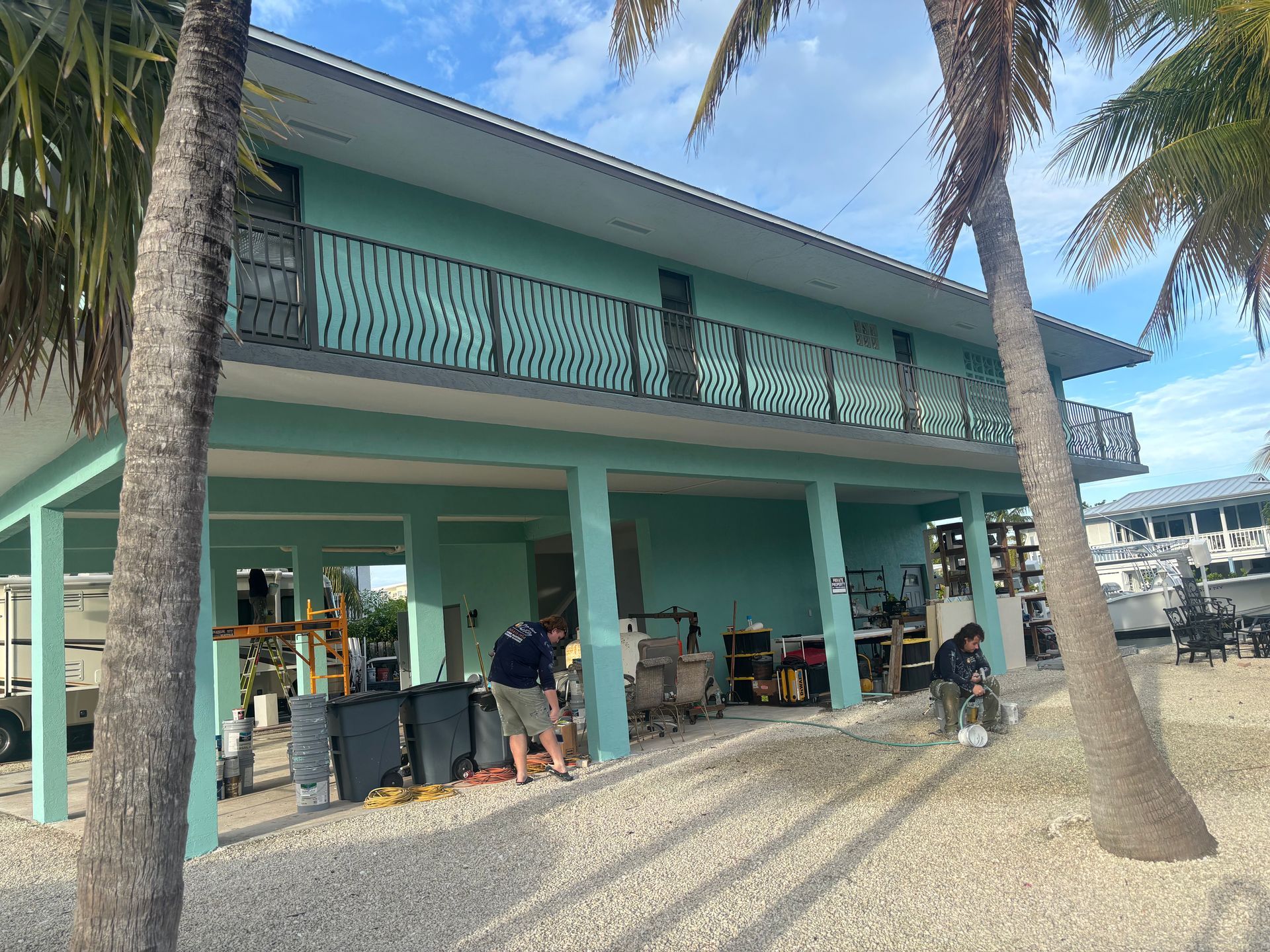 Turquoise beachside building with palm trees, balcony, and people gathered under the shaded patio