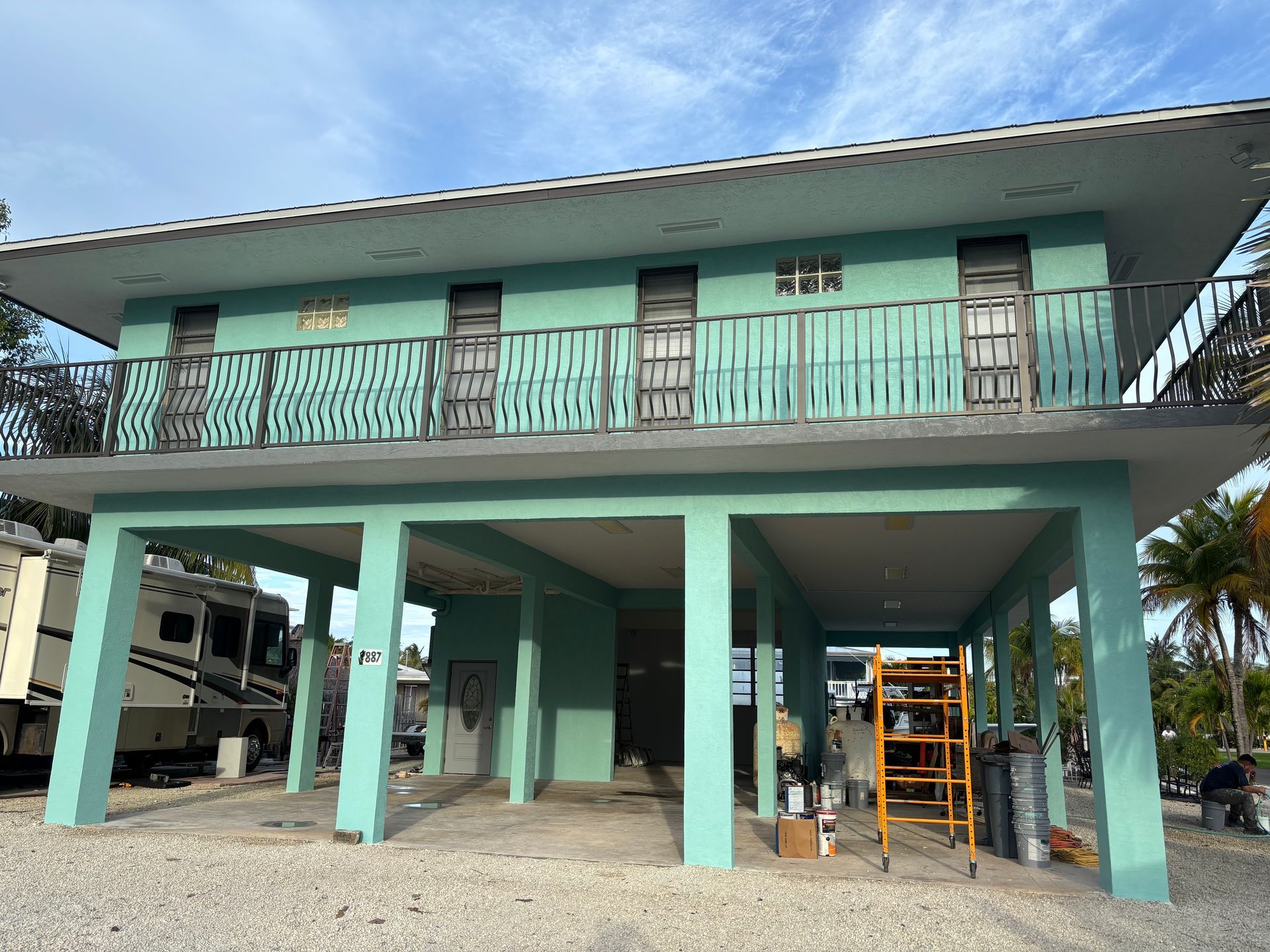Mint-green elevated house on stilts with a covered ground level and scaffolding nearby under a blue sky