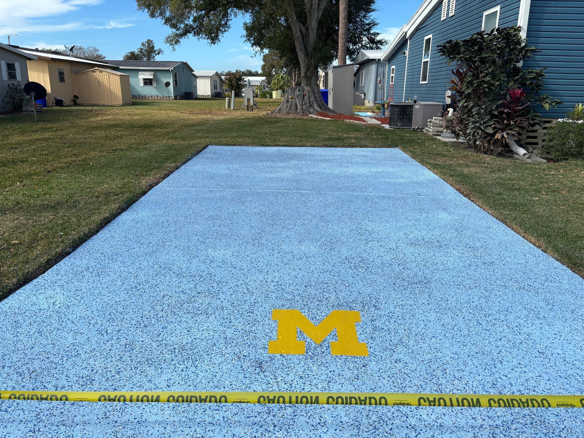Blue outdoor carpet with a yellow “M” logo on a walkway beside a house and lawn.