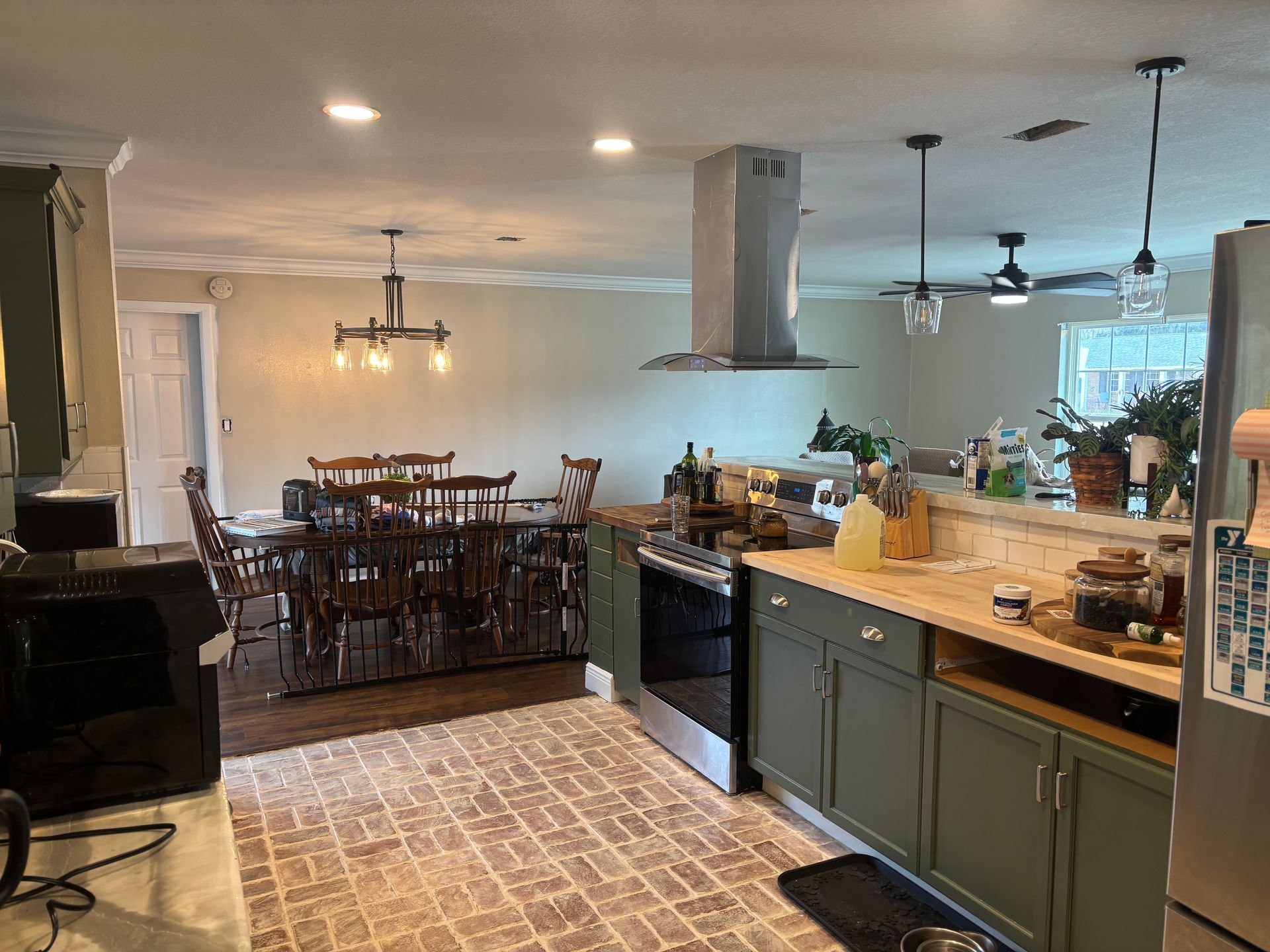 Open kitchen with dark green cabinets, brick floor, island, and dining area with wooden table and chairs