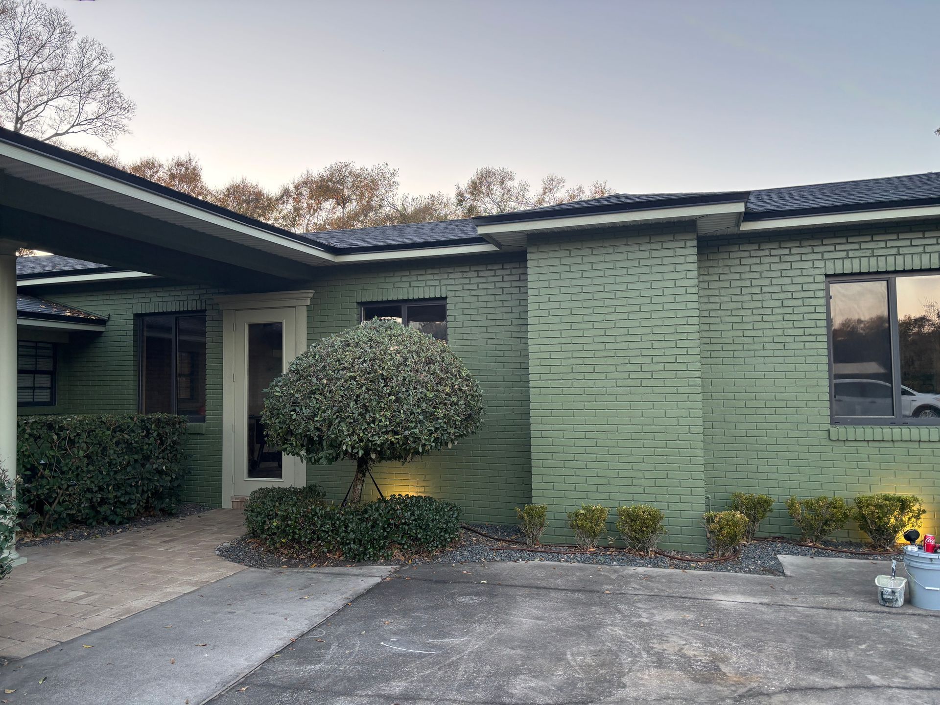 Front view of a green house with a carport, shrubs, and a driveway at dusk