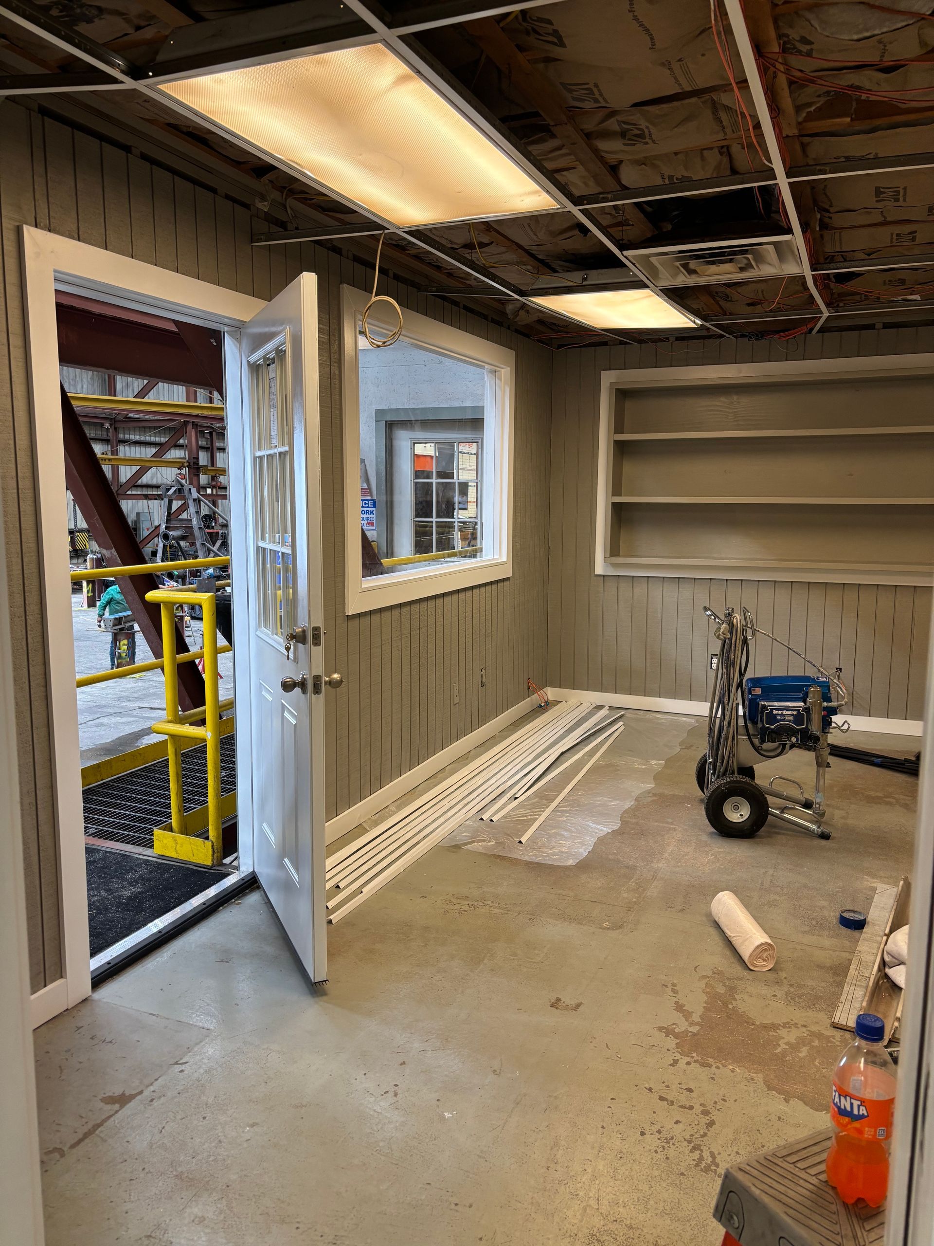 Interior room with open door, window, shelves, and painting equipment; gray walls, unfinished ceiling.