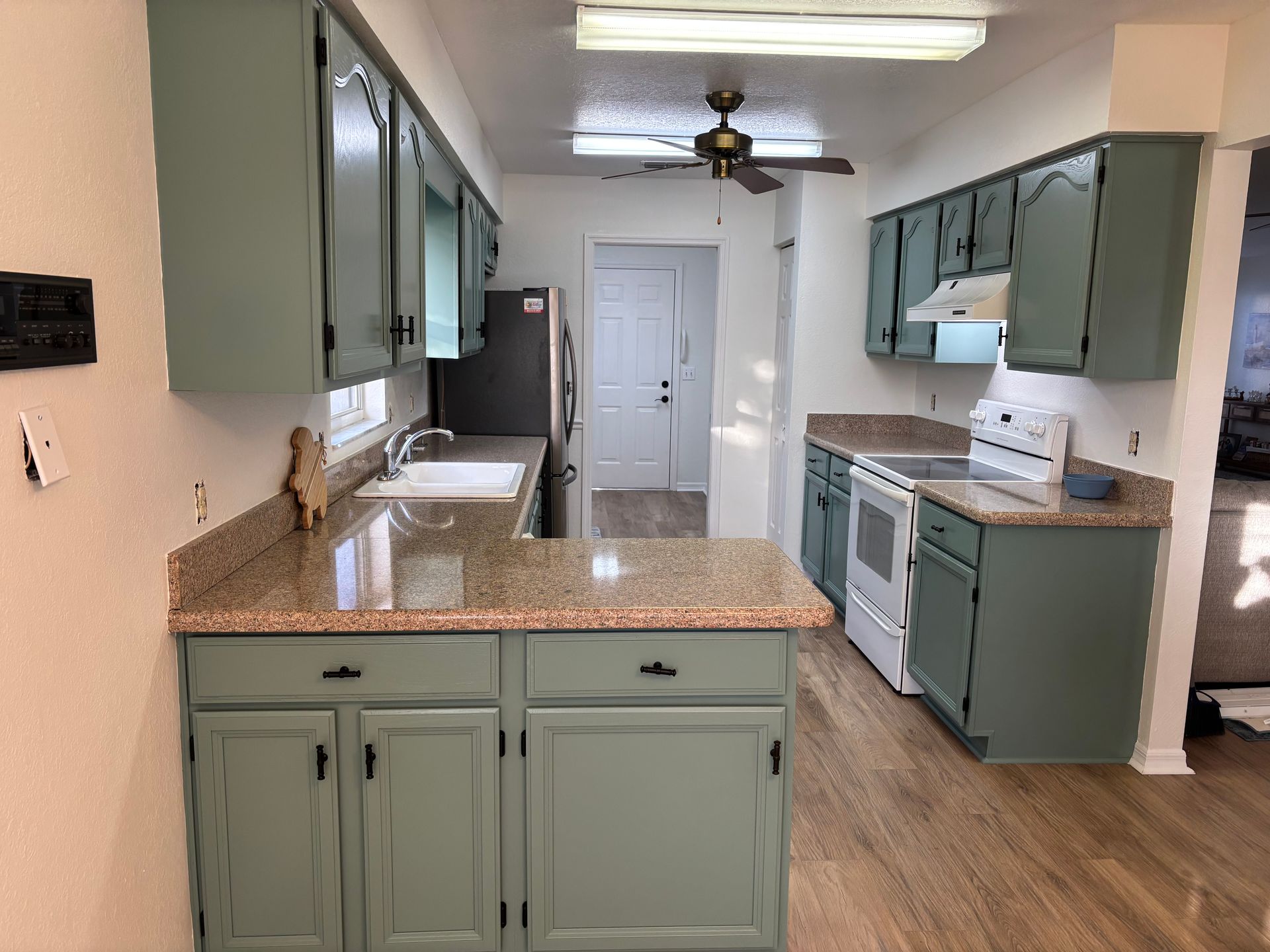 Kitchen with sage green cabinets, speckled granite island, wood floors, and stainless steel appliances.